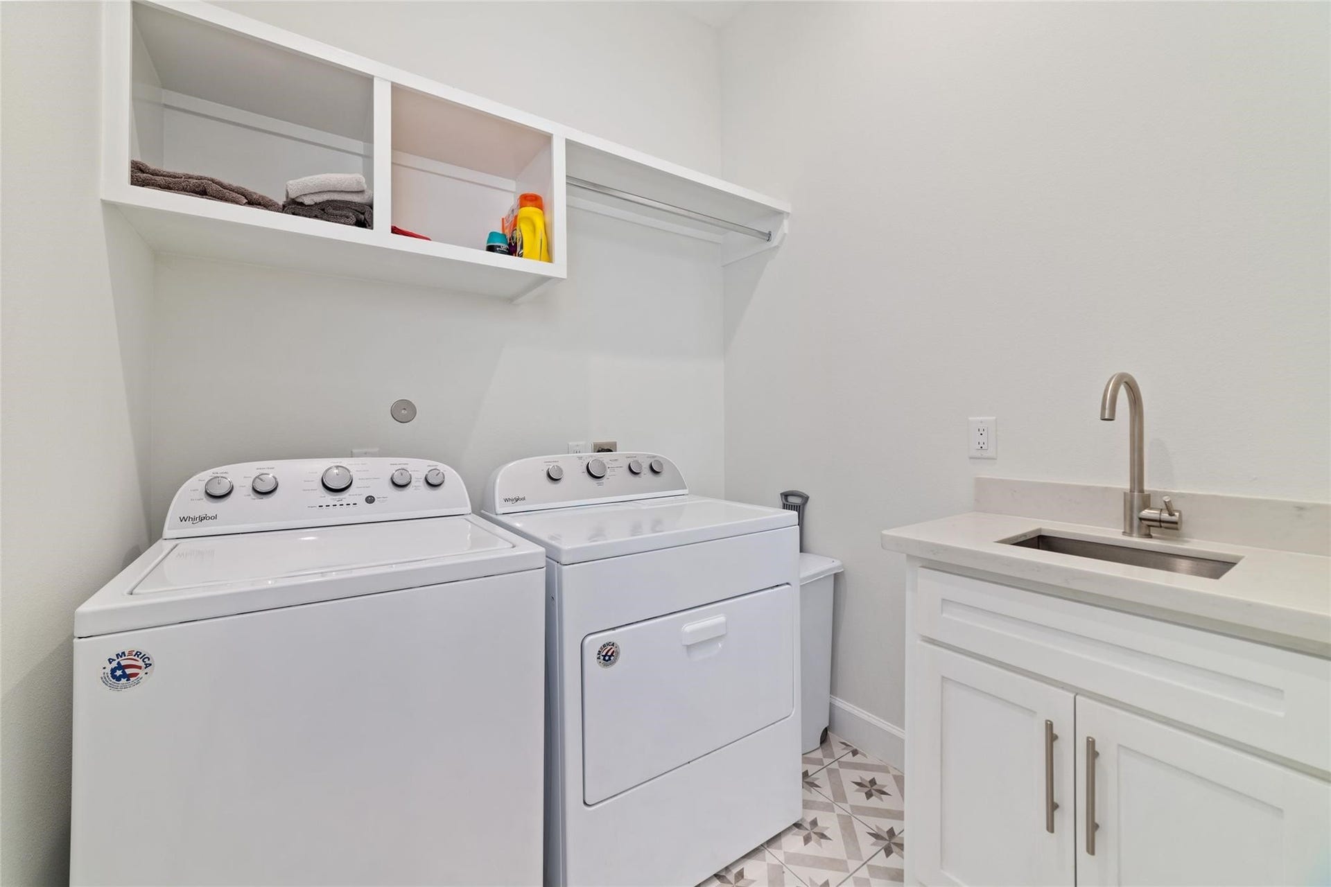 This is a bright, modern laundry room featuring a washer and dryer set, a sink with a countertop, and open shelving for storage. The patterned tile floor adds a stylish touch.