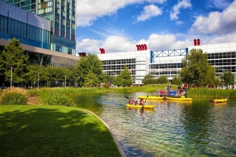 Discovery Green showcases a modern cityscape with a scenic pond where people are kayaking. Surrounded by lush greenery and contemporary buildings, it offers a blend of urban living and outdoor recreation.