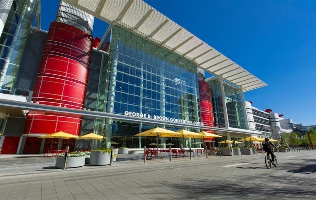 George R. Brown Convention Center with striking red cylindrical structures and expansive glass facades. The area is spacious with wide walkways and outdoor seating under yellow umbrellas, a bustling urban environment nearby.