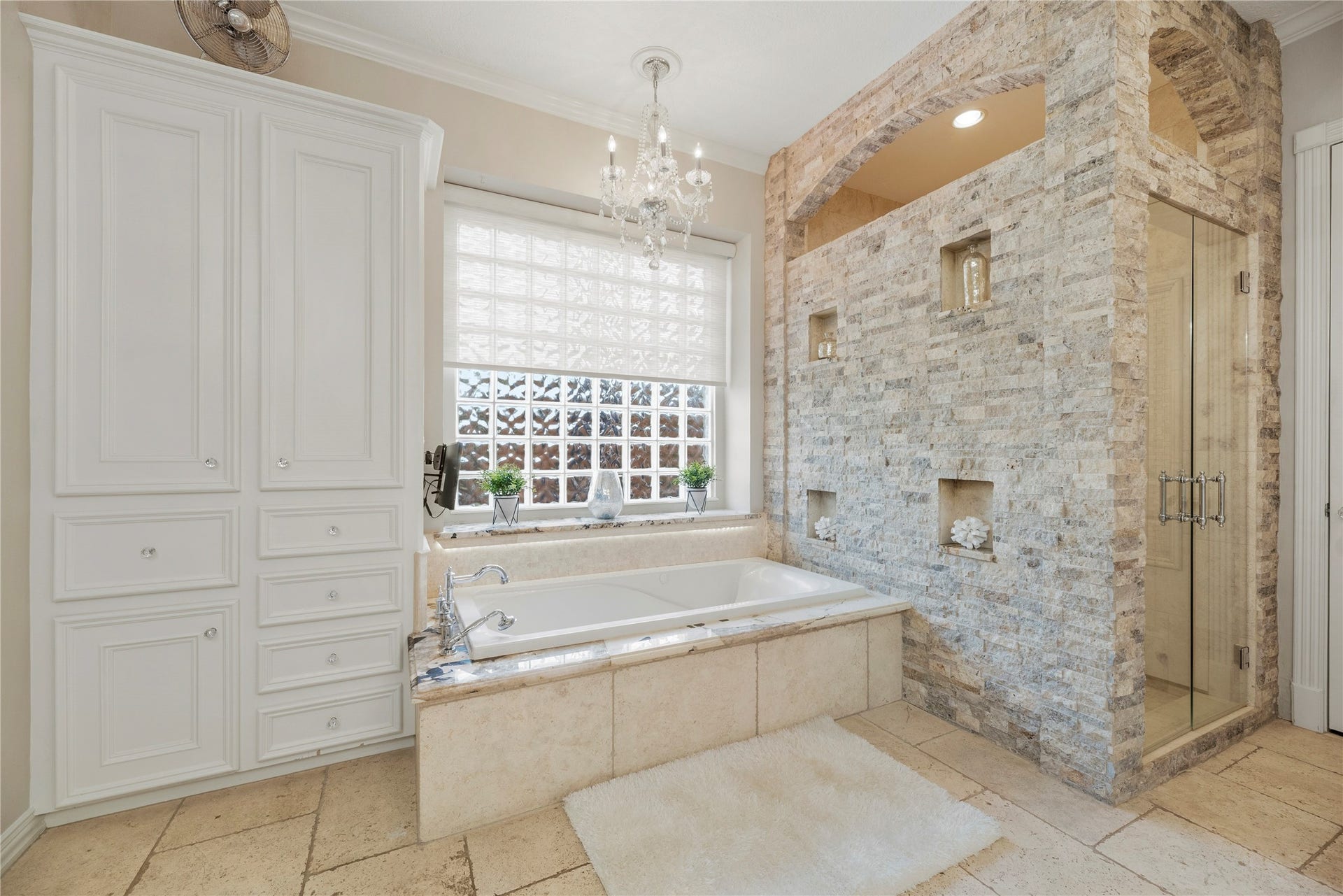 13915 Springmint Drive, Cypress, TX 77429 Luxurious primary bathroom features a stunning stone-clad walk-in shower with architectural arches and decorative niches, a generous jetted tub beneath a glass block window, elegant chandelier and extensive custom cabinetry.