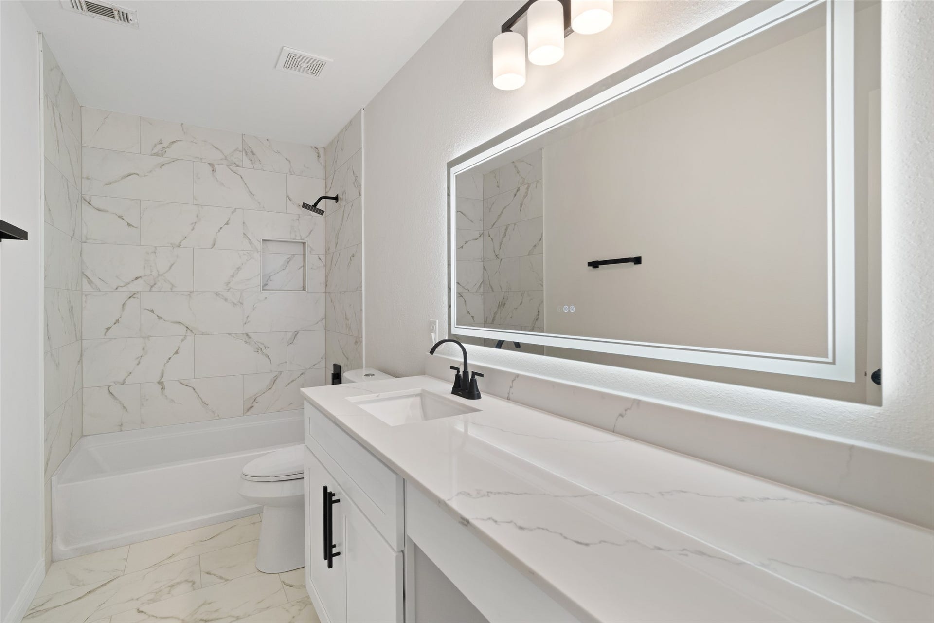 21611 Manitou Falls Lane, Katy, TX 77449 Full Bathroom Updated bathroom featuring a long vanity with stone-look countertop, illuminated mirror, matte-black fixtures, and a tub/shower surrounded by marble-look tile.