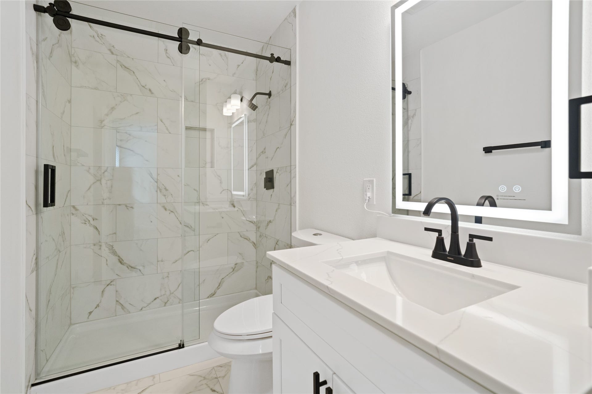 21611 Manitou Falls Lane, Katy, TX 77449 Full Bathroom Stylish bathroom with marble-look shower tile, a glass sliding enclosure, stone-look vanity top, illuminated mirror, and contrasting matte-black fixtures.