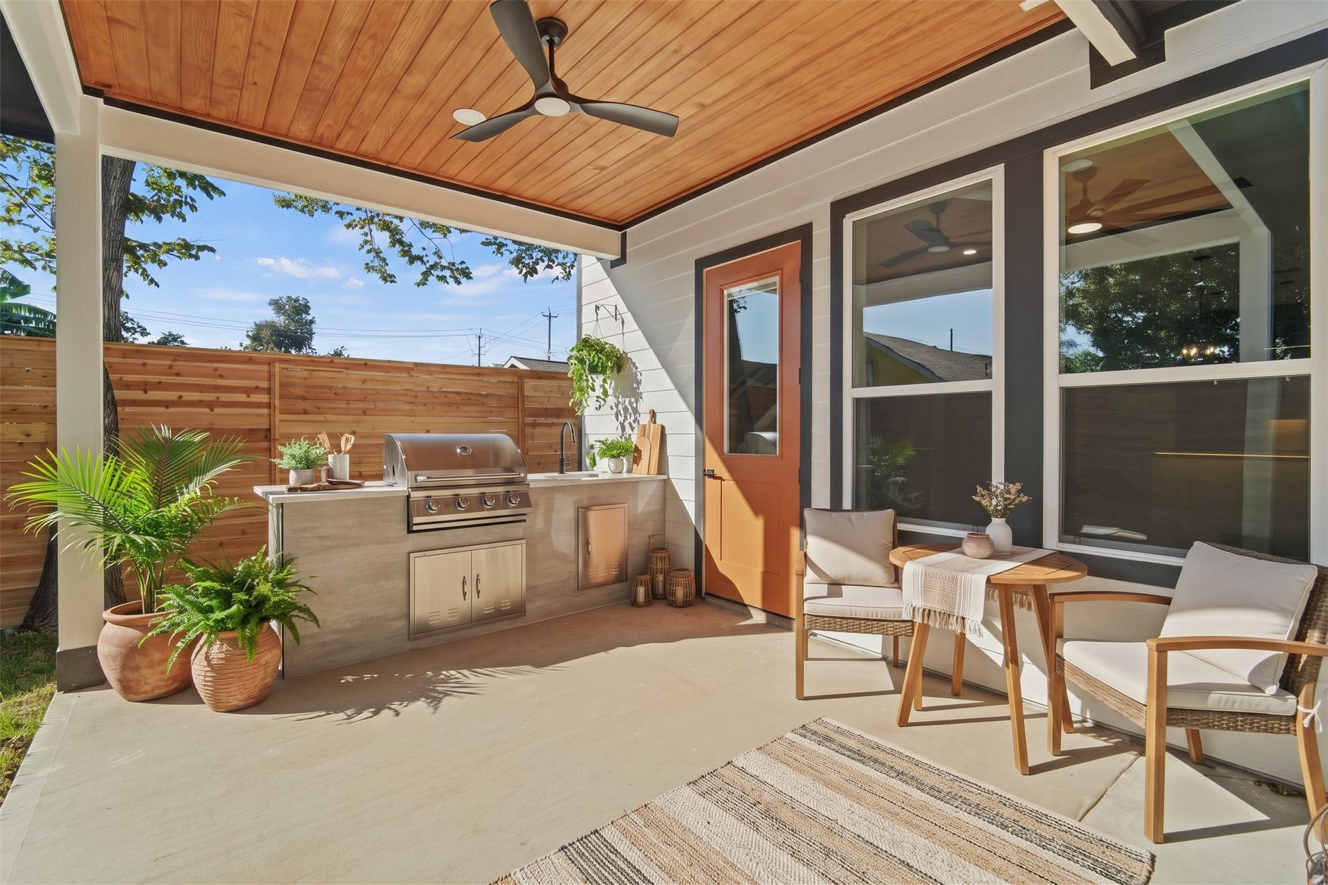 Virtually Staged: This covered back patio features a built-in outdoor kitchen island equipped with stainless steel storage drawers, a sink, and prep counter space. A natural wood ceiling with a modern fan anchors the sheltered space.