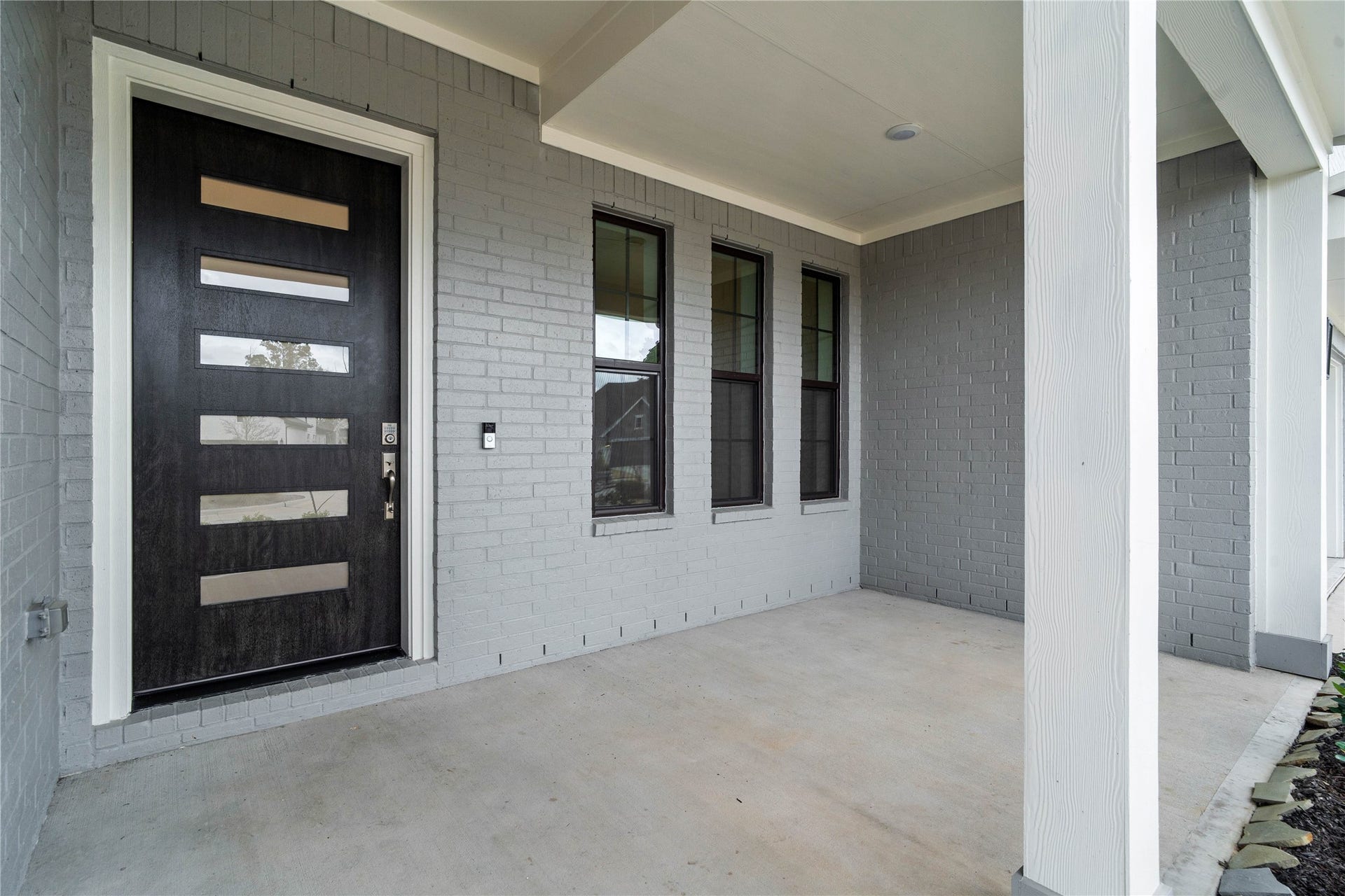 This covered front porch offers a clean, contemporary feel with its sleek grey brick faade and smooth concrete flooring. The space is anchored by a stunning modern five-lite glass front door in a rich dark finish, which provides an inviting and sophisticated entrance to the home.