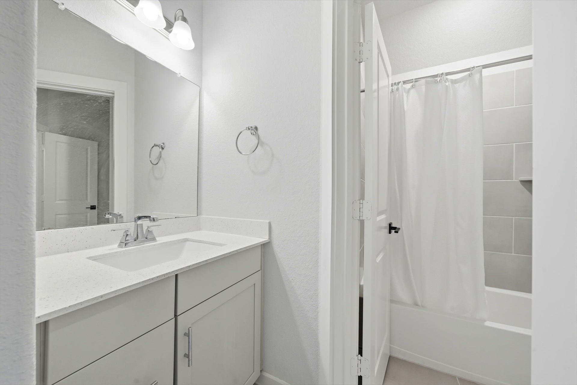 8815 Morning Glow Drive, Missouri City, TX 77459 This photo shows a modern, bright bathroom featuring a sleek white vanity with a quartz countertop, a large mirror, and stylish lighting. There's a shower-tub combo with a tiled surround and a white shower curtain, offering a clean and inviting space.