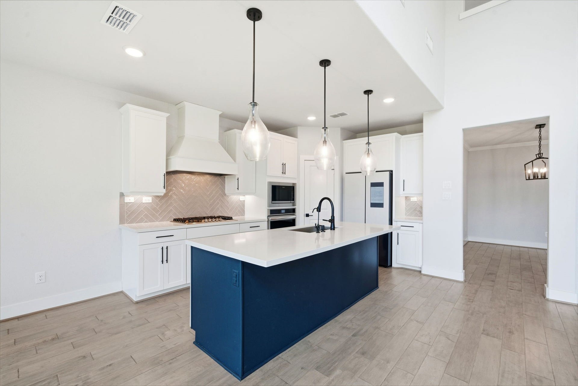 8815 Morning Glow Drive, Missouri City, TX 77459 This modern kitchen features a spacious island with a sleek navy base and white countertop, pendant lighting, and white cabinetry. It includes a built-in oven, gas stovetop, and contemporary fixtures, all set on light wood flooring, creating a bright and inviting space.