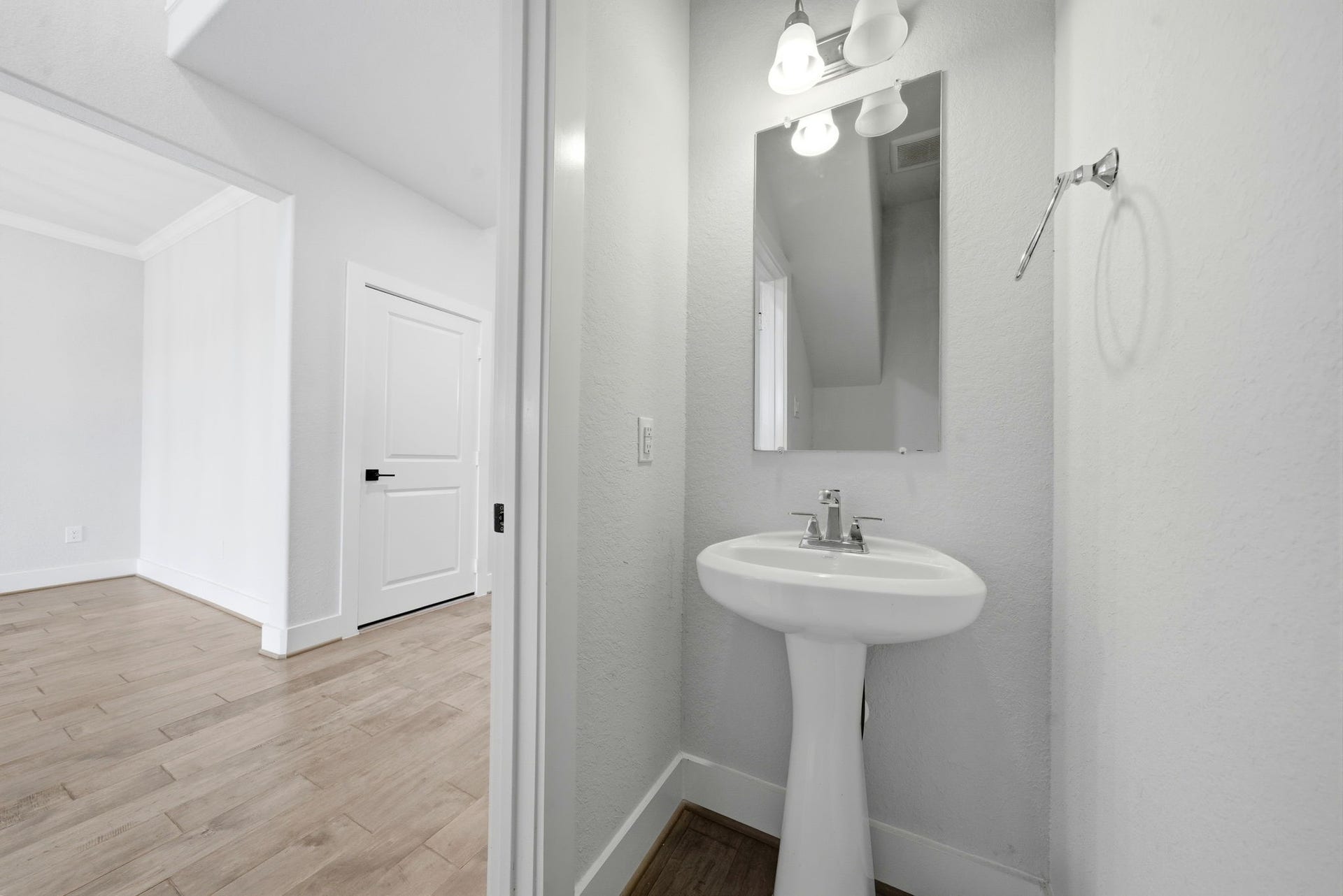 8815 Morning Glow Drive, Missouri City, TX 77459 This photo shows a compact, modern powder room with a pedestal sink, large mirror, and stylish lighting. Adjacent is a bright space with light wood flooring and a white door, adding to the home's clean and contemporary aesthetic.