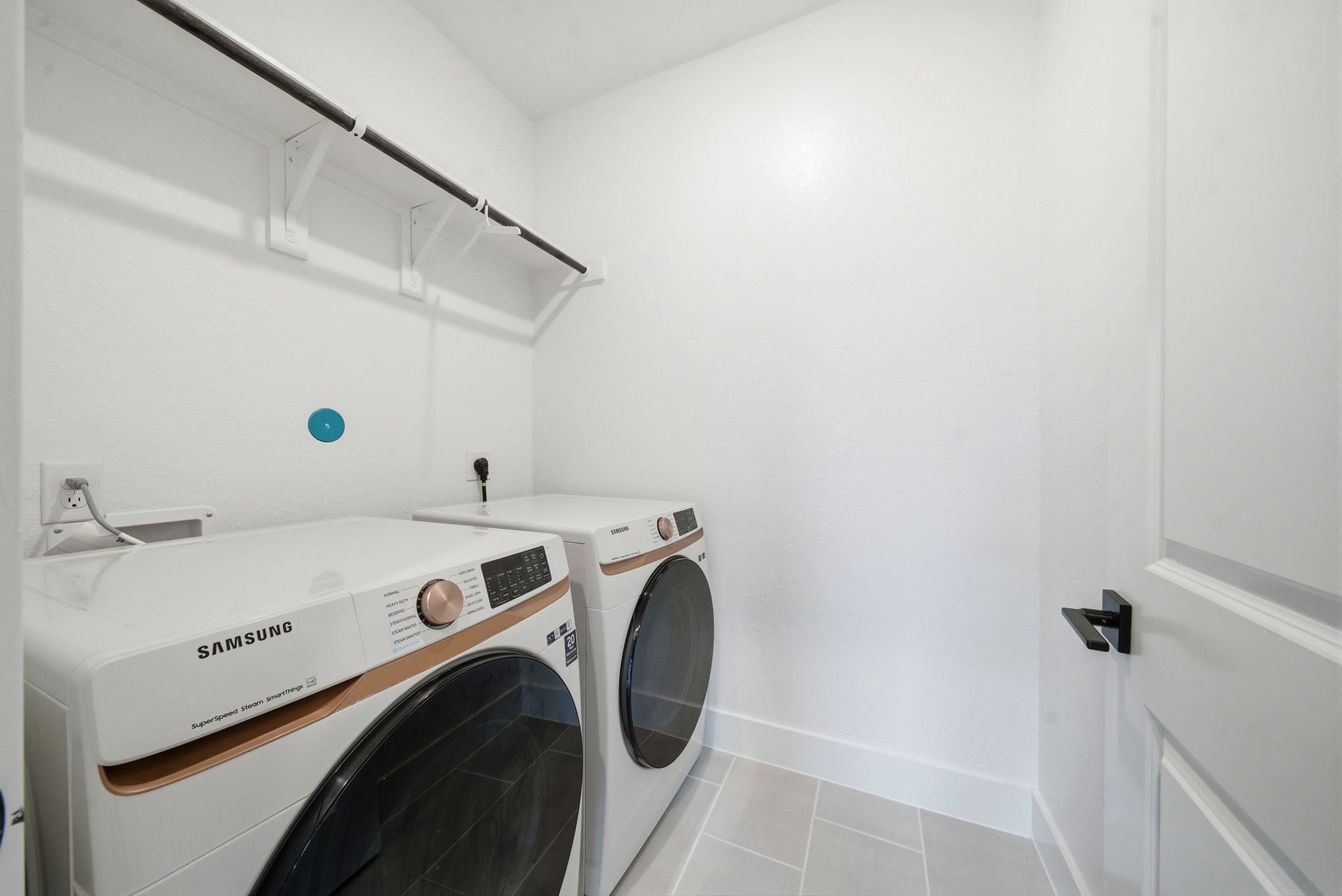 8815 Morning Glow Drive, Missouri City, TX 77459 This is a modern laundry room featuring a Samsung washer and dryer set. The space is bright with white walls, light tile flooring, and includes a convenient shelving rack above the appliances for storage.