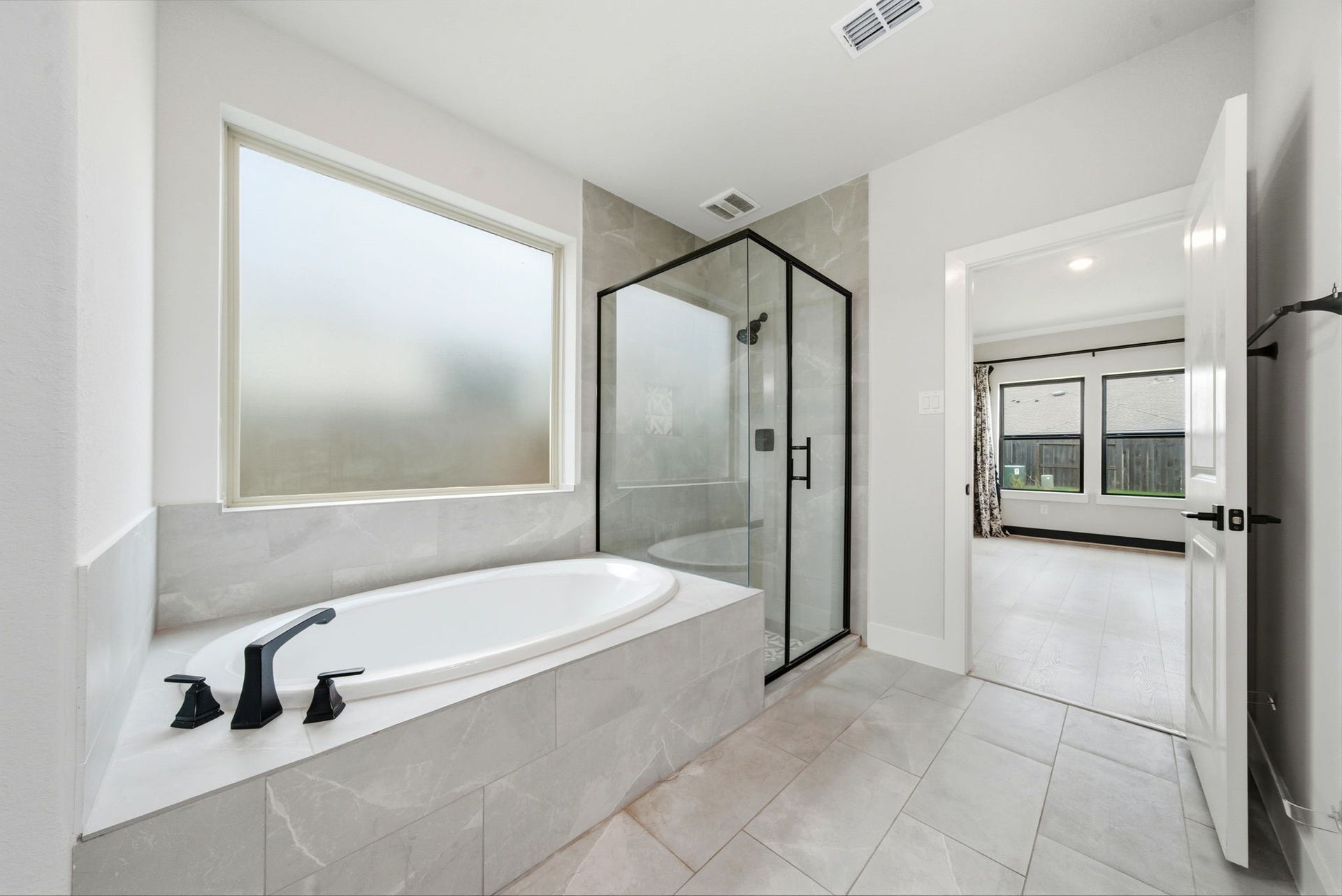8815 Morning Glow Drive, Missouri City, TX 77459 This bathroom features a spacious design with a large soaking tub, a sleek glass-enclosed shower, and bright natural light from a frosted window. It leads directly into a bright adjoining room with large windows.