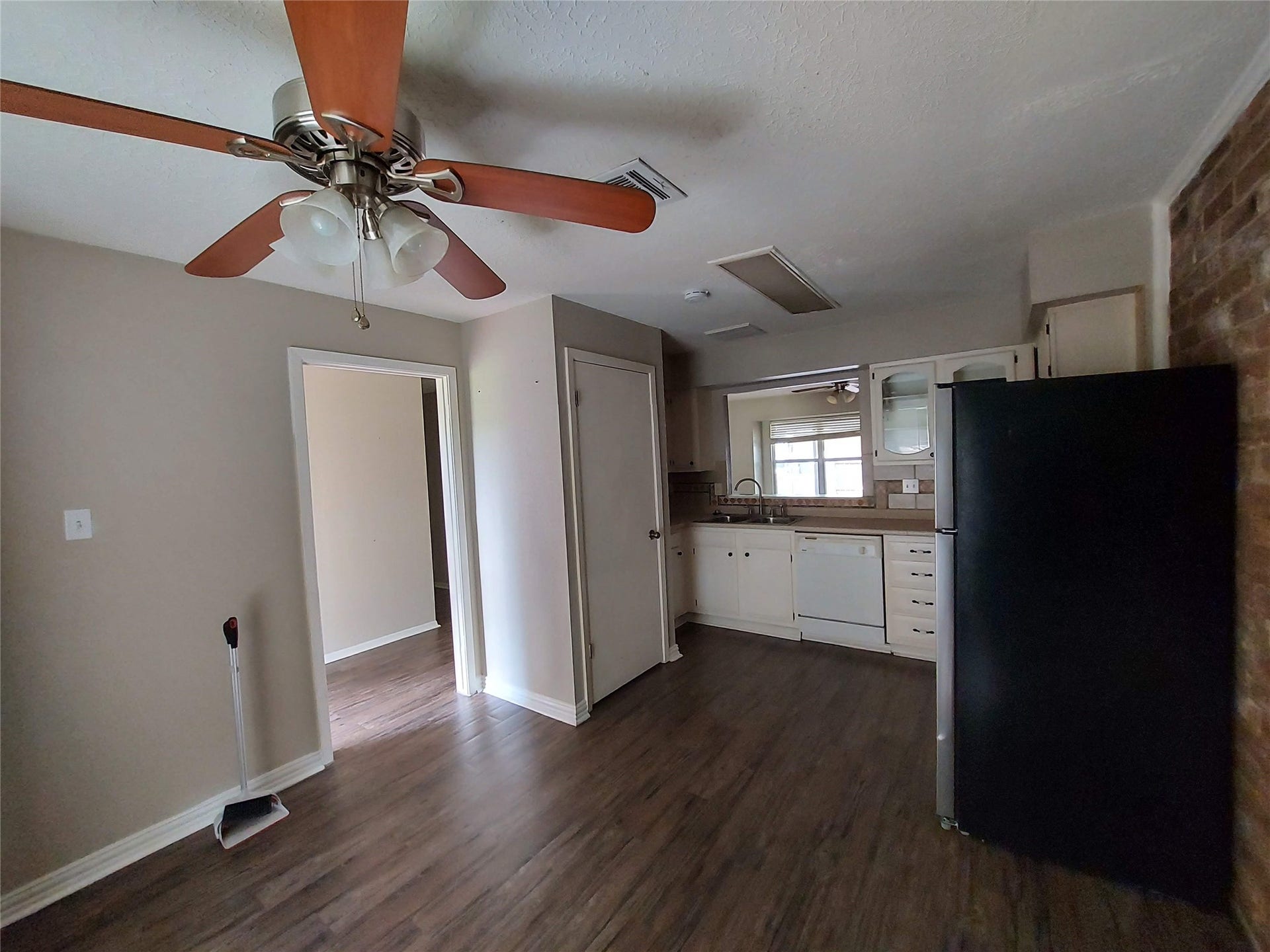 3878 Laura Leigh Drive, Friendswood, TX 77546 Cozy kitchen with warm wood flooring, a ceiling fan, and a charming brick accent wall. The space features ample cabinetry, a sleek black refrigerator, and a convenient pass-through window to the living area.