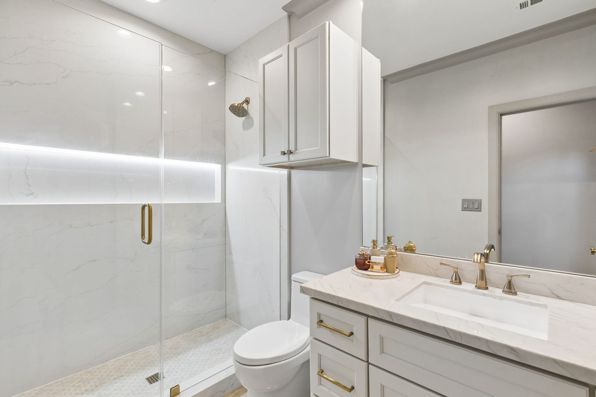 The secondary bathroom measures approximately 5'6" x 9'9" and is beautifully appointed with upscale finishes including Quartzite countertop, shaker-style cabinetry, and brushed brass hardware create a timeless design. A frameless marble shower with Marble surround and lighted shower niche and additional storage cabinet enhance both style and functionality.