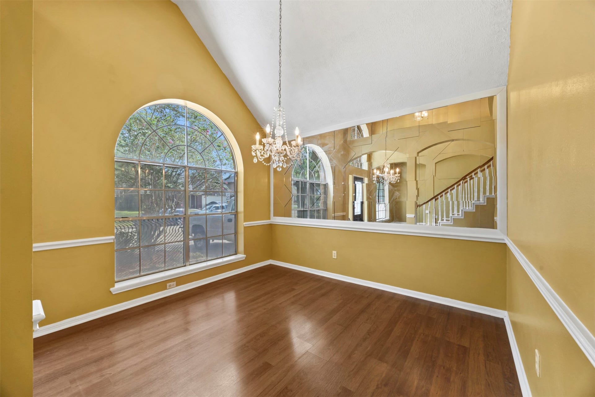 19119 Timber Trace Drive, Humble, TX 77346 Elegant dining room featuring high vaulted ceilings, a striking chandelier, and a large arched window that fills the space with natural light, complemented by warm wood flooring and tasteful wainscoting.