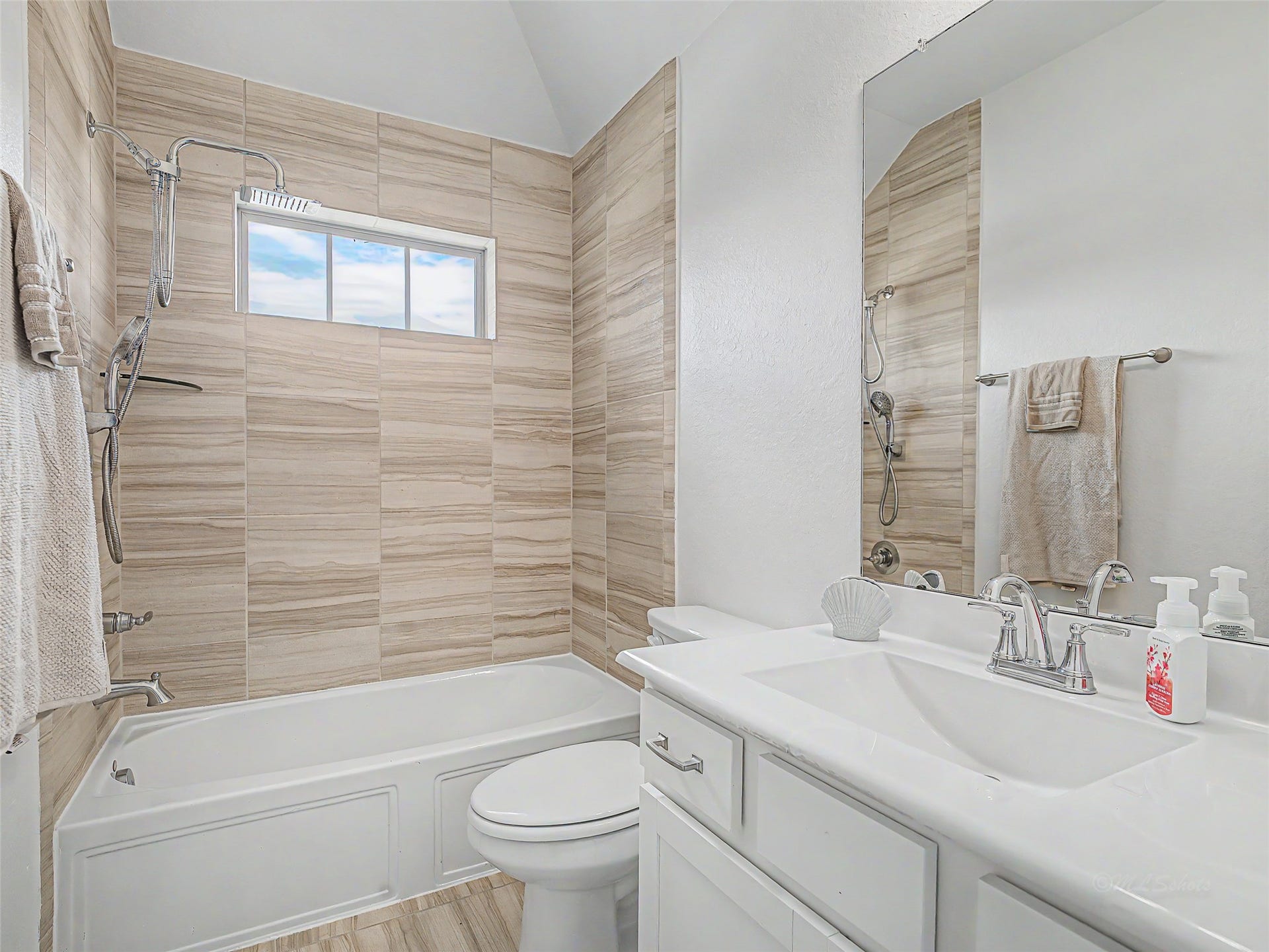 6019 Dovetail Cliff Court, Katy, TX 77493 The combination shower and soaking tub is enclosed by full-height, travertine-style ceramic tile surround in warm neutral tones, extending all the way up to the vaulted ceiling line. A high transom window above the tub allows natural light to stream in while maintaining complete privacy.