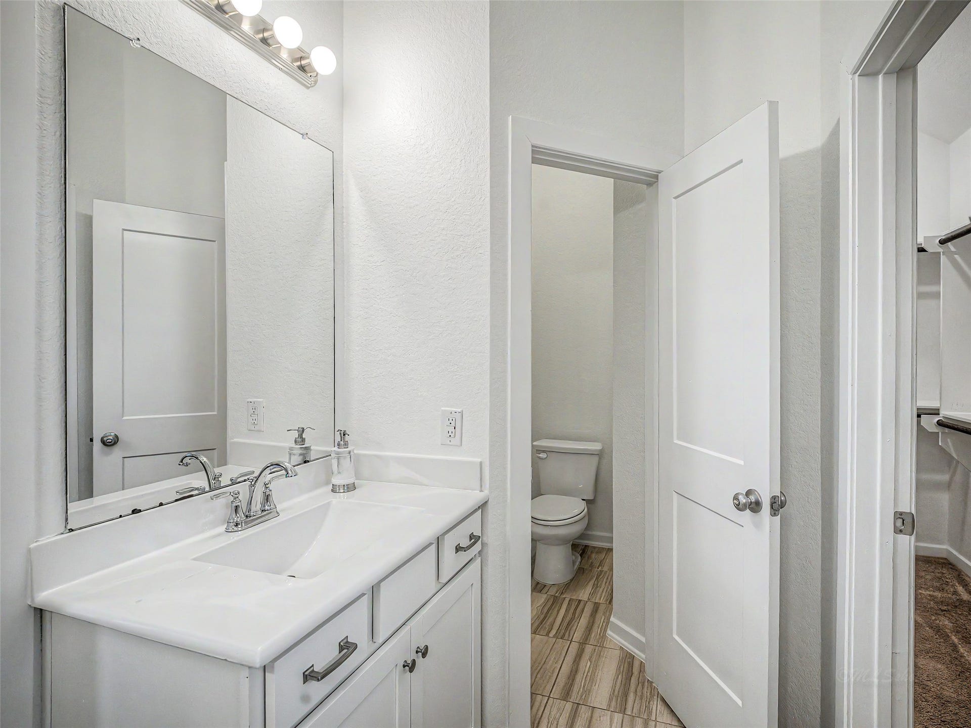 6019 Dovetail Cliff Court, Katy, TX 77493 This en-suite primary bathroom features a long white vanity with an integrated sink, brushed chrome hardware, a vast frameless mirror, and a multi-bulb vanity light bar. A private water closet with a swinging door isolates the toilet, while tile flooring coordinates with the overall neutral finish.