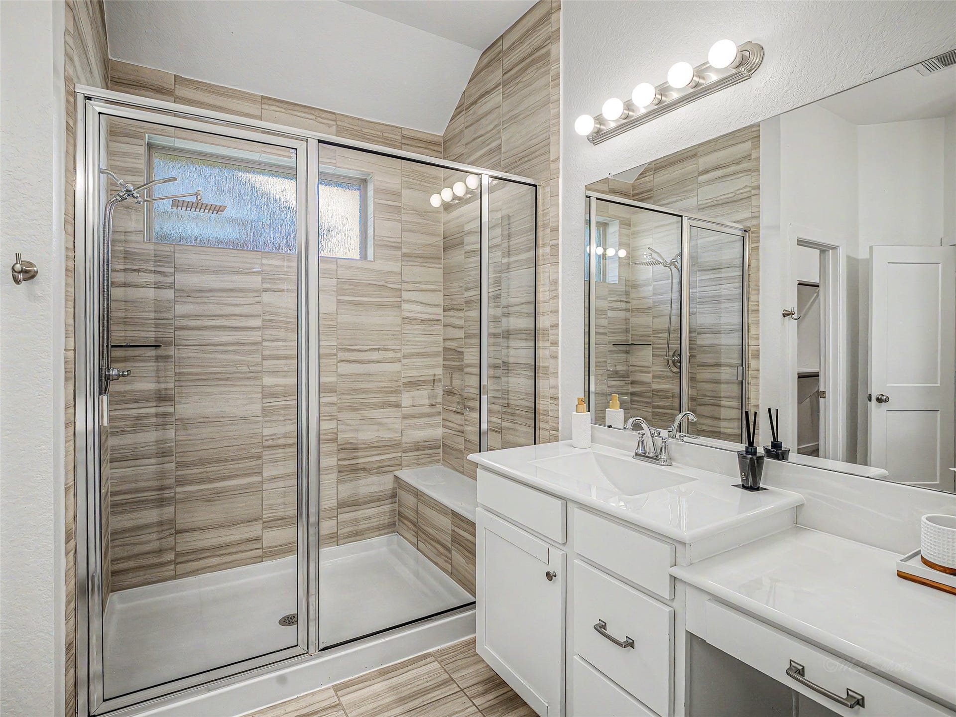 6019 Dovetail Cliff Court, Katy, TX 77493 This bathroom features a walk-in glass shower with a full-height, travertine-style tile surround, built-in tile bench, overhead rainfall showerhead, handheld sprayer, and a high frosted-glass transom window for natural light and privacy.