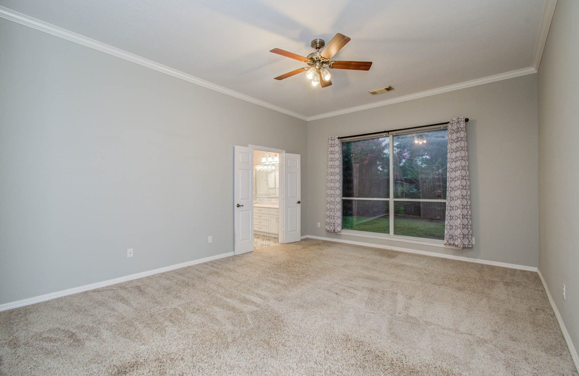 14403 Red Tailed Hawk Lane, Houston, TX 77044 Spacious bedroom with plush carpeting, featuring a large window with elegant drapes offering plenty of natural light and a ceiling fan for comfort. An en-suite bathroom with a dual vanity is accessed through double doors.