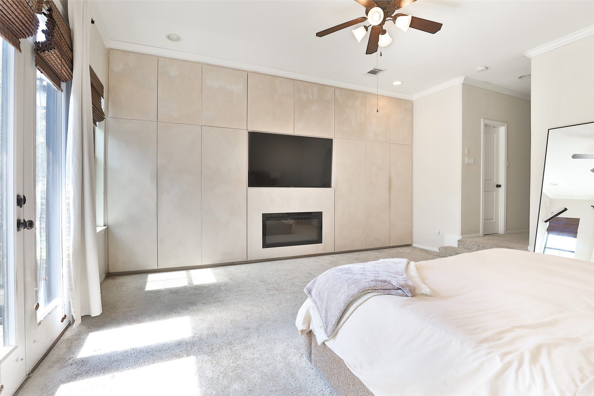 1112 Bingham Street, Houston, TX 77007 Elegant bedroom with a built-in entertainment wall featuring a sleek TV and modern fireplace, complemented by natural light streaming through large windows and French doors.