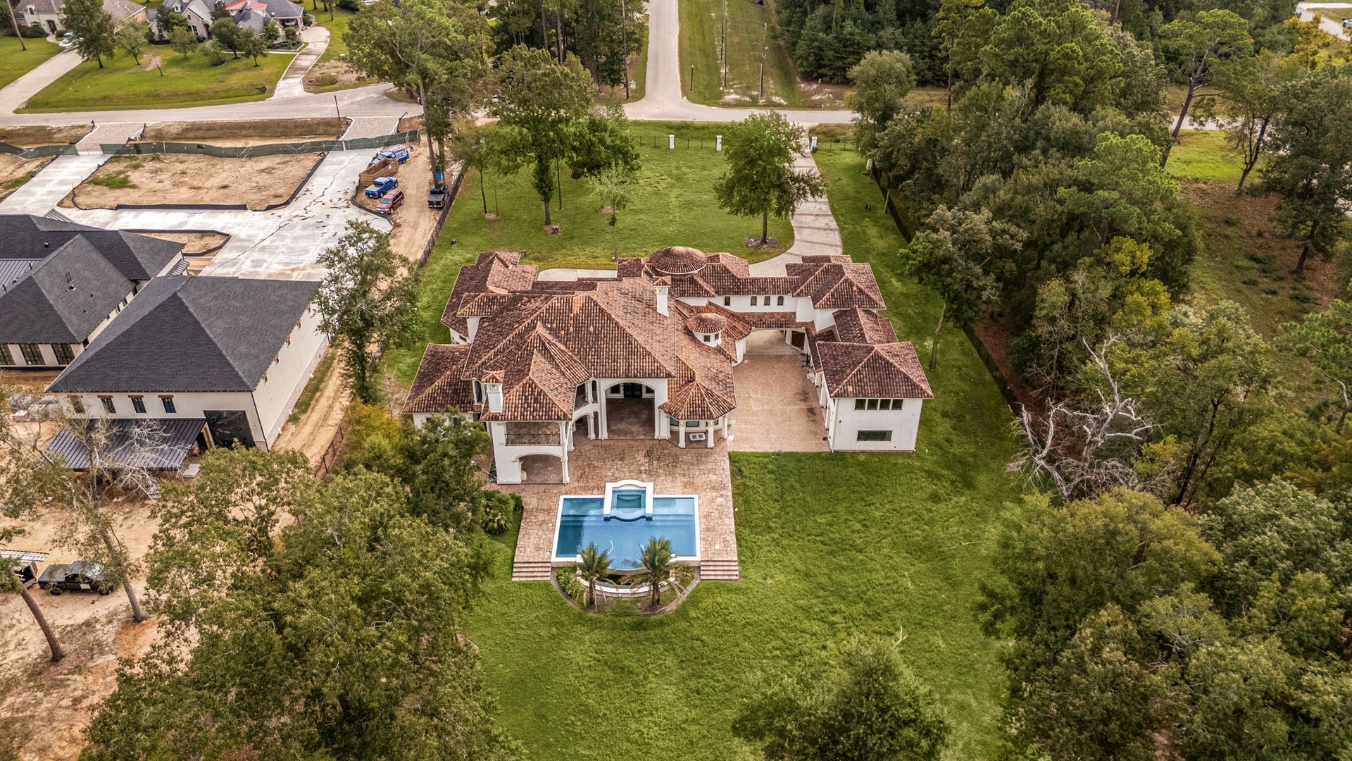 This stunning overhead aerial view showcases the full layout of the magnificent resort-style estate. Nestled perfectly among dense, mature trees, the property highlights its expansive green backyard, the sparkling blue swimming pool with its integrated spa, and the massive brick-paver motor court layout. This perspective beautifully emphasizes the balance of private luxury, expansive acreage, and timeless architectural design.