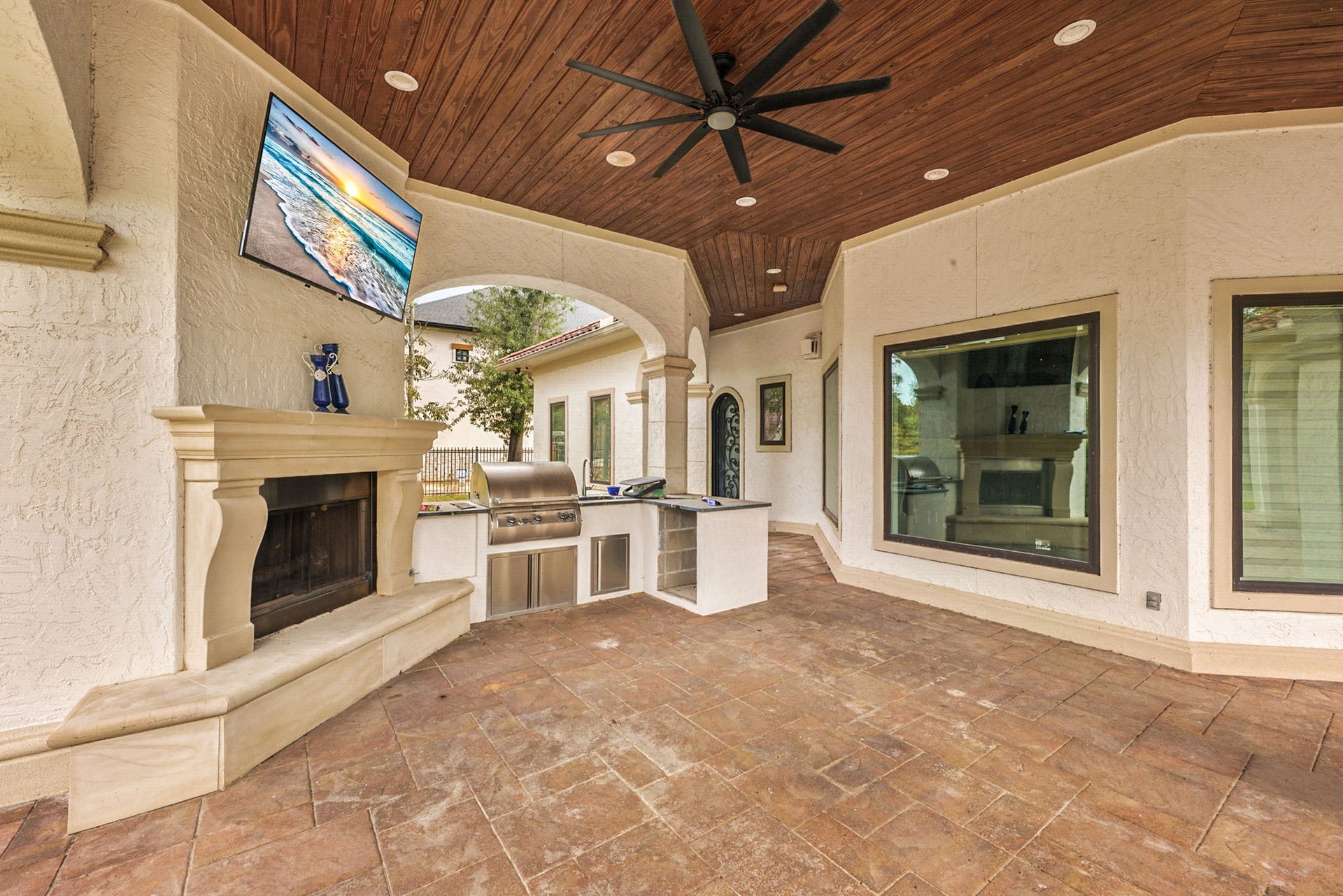 Experience ultimate outdoor entertaining in this sprawling covered patio oasis. The space features a warm wood-plank ceiling with a large fan, recessed lighting, and elegant flagstone flooring. It comes fully equipped with a magnificent stone-mantel fireplace, a mounted television setup, and a complete outdoor summer kitchen for hosting guests in style.