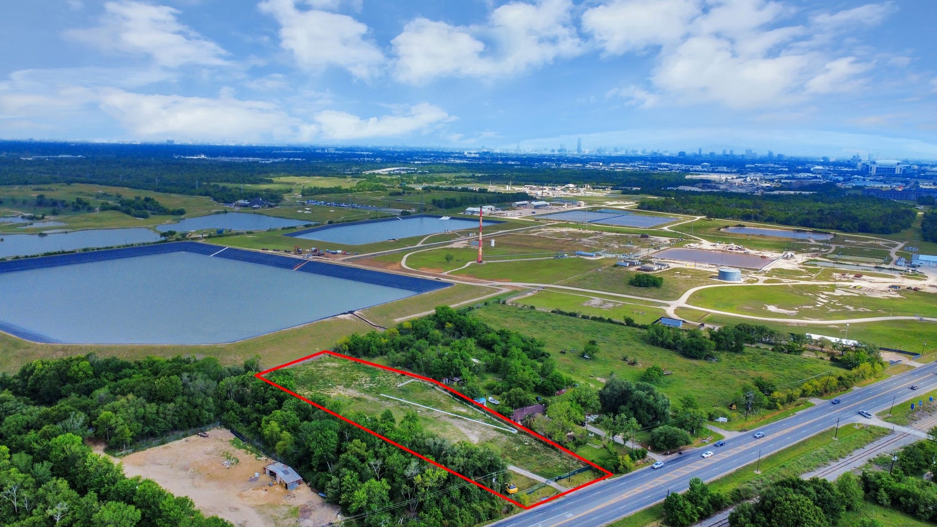 11826 Almeda Road, Houston, TX 77045 This aerial photo showcases a large plot of land outlined in red, situated near a major road. The area is surrounded by greenery and open fields, with industrial facilities and water bodies visible in the distance. The skyline of a city is faintly visible on the horizon, suggesting a mix of rural and urban proximity.