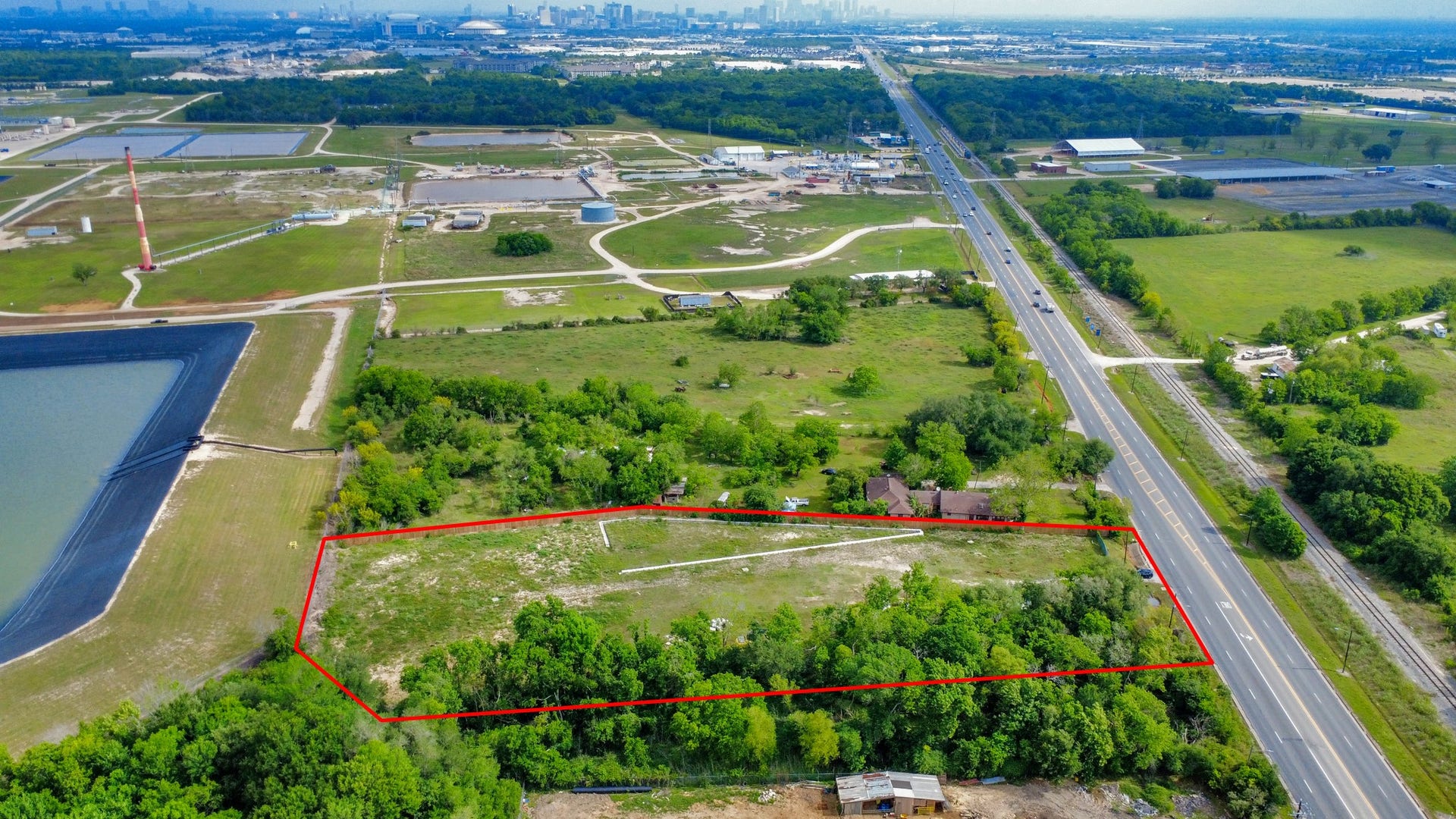11826 Almeda Road, Houston, TX 77045 This aerial photo showcases a spacious, rectangular plot of land outlined in red, bordered by greenery. It's situated near a major road with easy access to urban amenities. The surrounding area features open fields and some industrial facilities, with the city skyline visible in the distance.