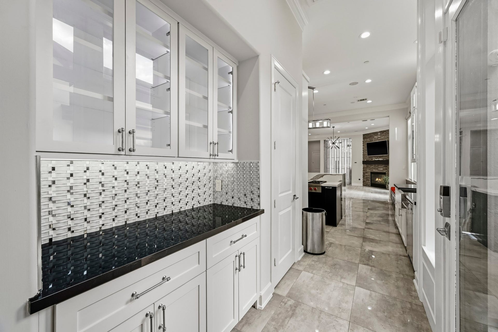 Elegant kitchen hallway featuring sleek black countertops and white cabinetry, highlighted by a modern mosaic backsplash. The open layout leads to a spacious living area with a cozy fireplace.