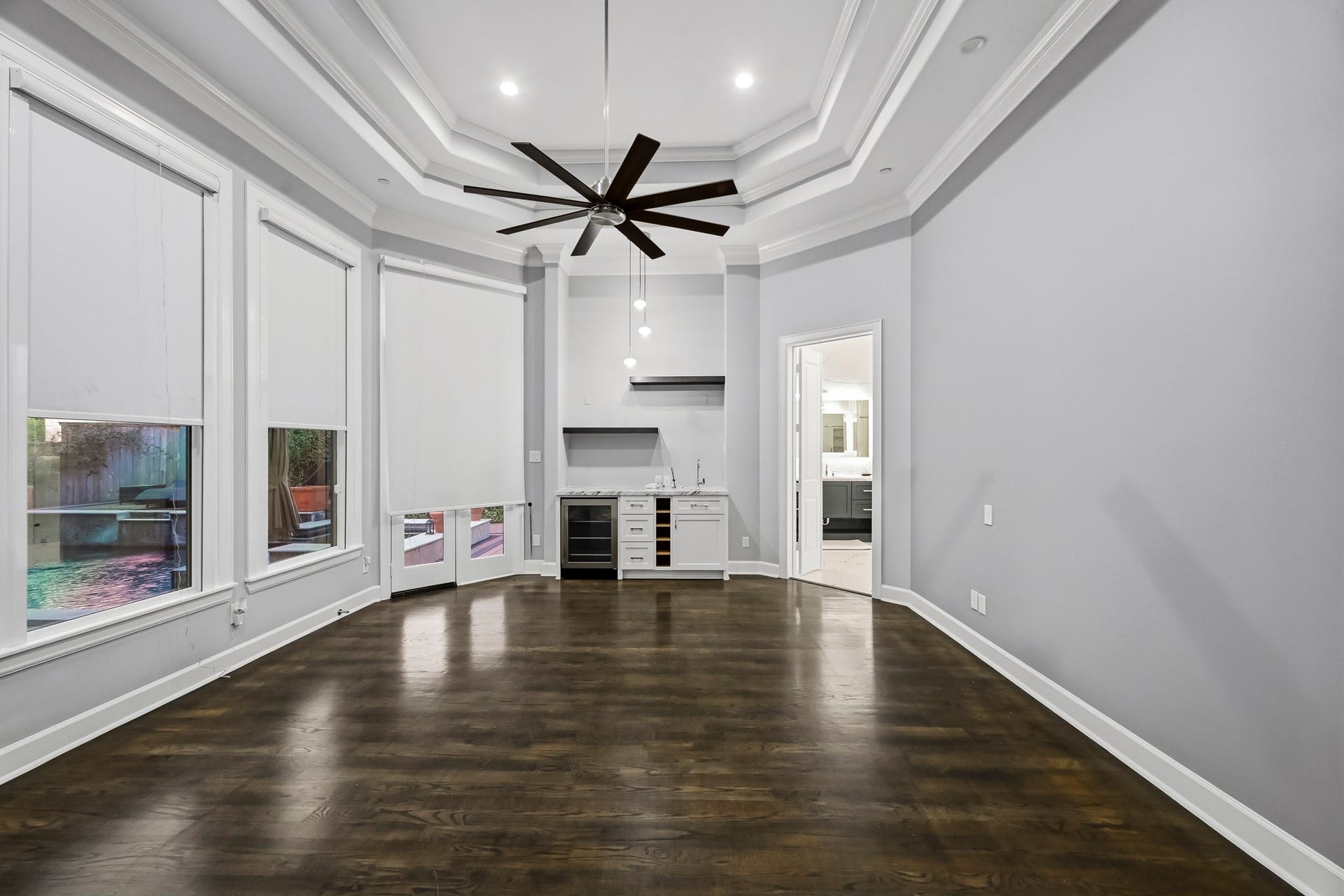 Elegant primary bed room featuring rich floors, a stylish tray ceiling with a modern fan, and ample natural light from large windows with shades. A convenient wet bar adds functionality, while the open layout provides seamless access to an adjacent space.