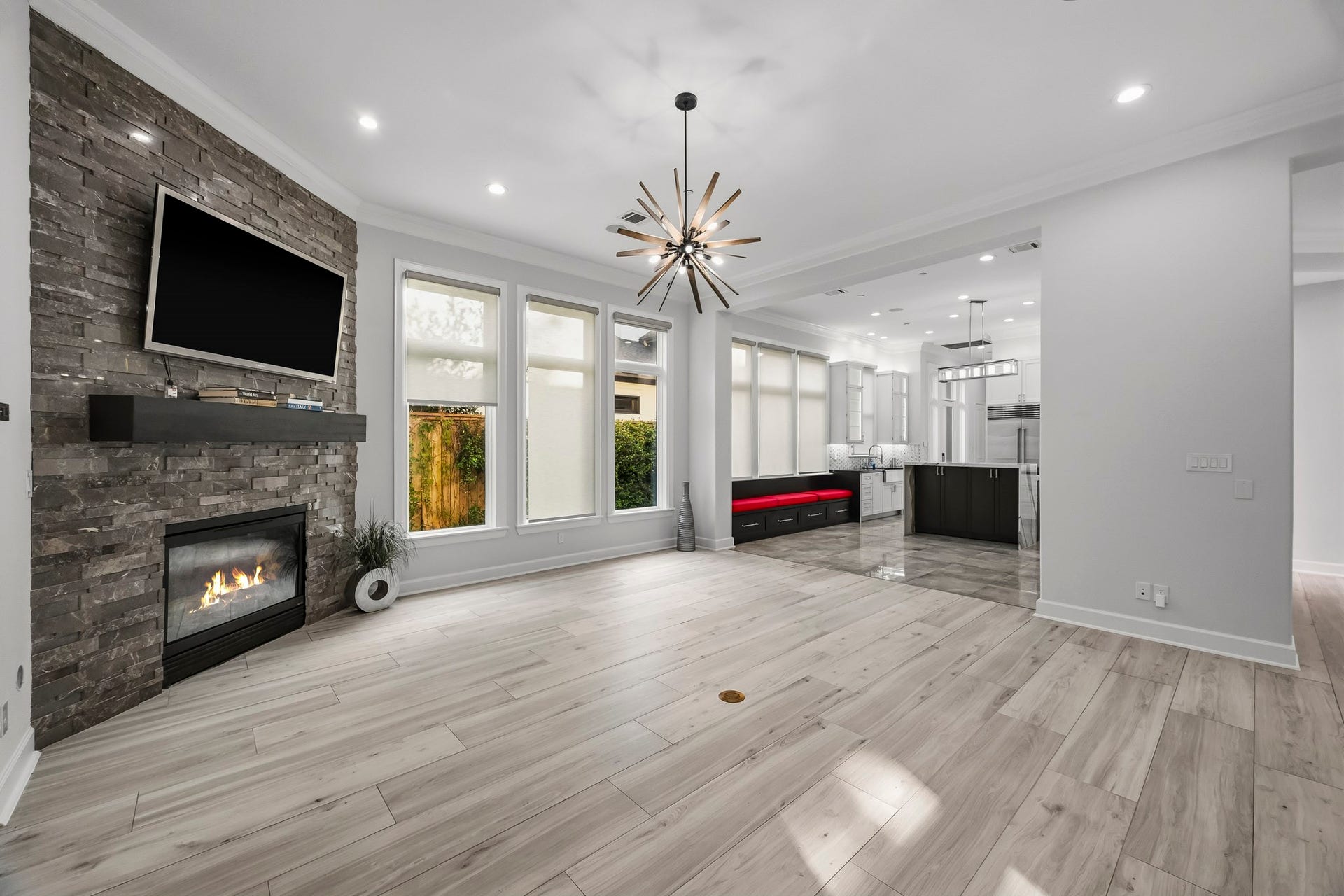 Spacious open-concept living area with a modern stone fireplace, elegant chandelier, and abundant natural light, seamlessly connecting to a sleek, contemporary kitchen.
