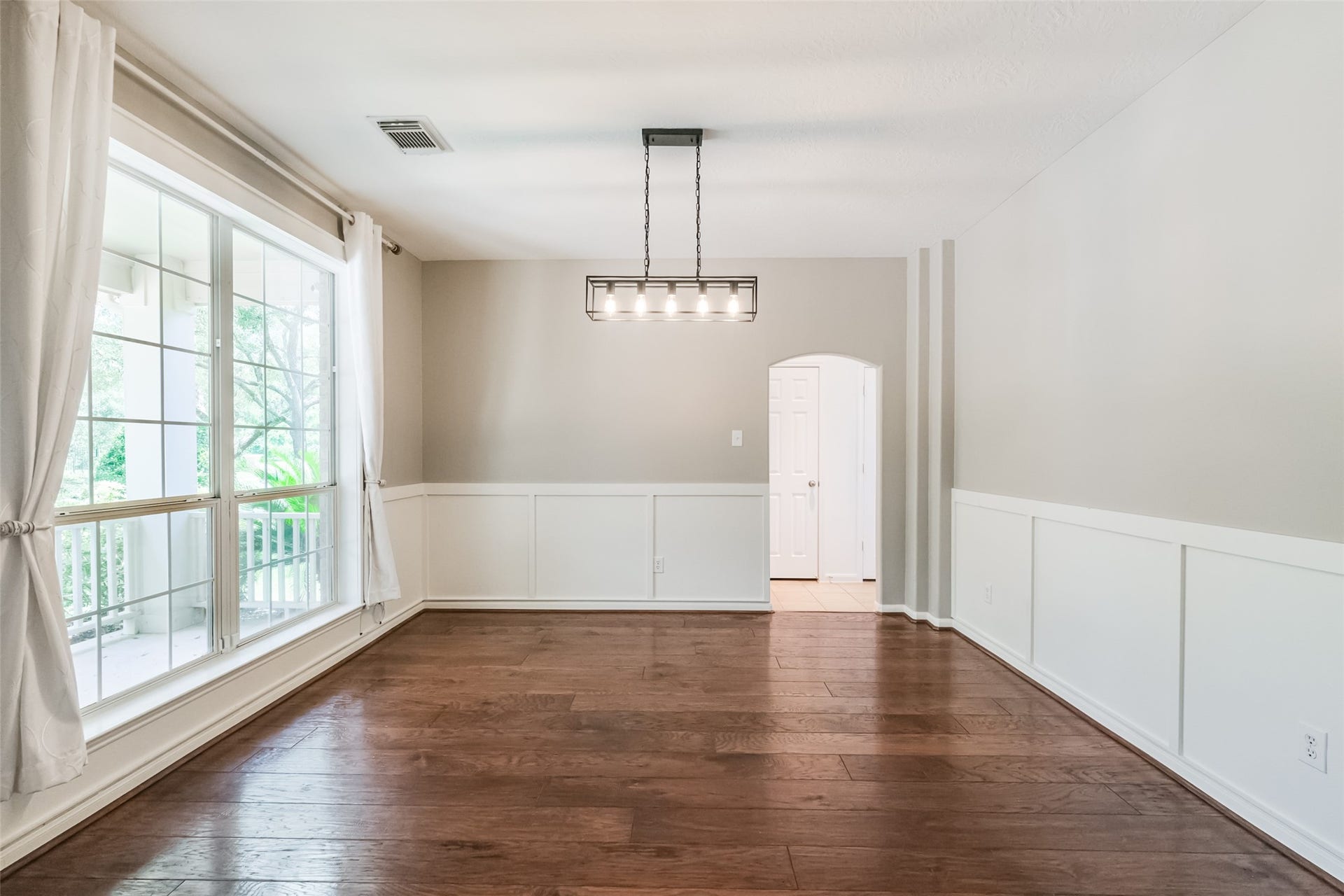 10314 Five Oaks Crossing, Missouri City, TX 77459 This bright dining room features large windows with white curtains, wood flooring, neutral walls with wainscoting, and a modern light fixture, offering a welcoming and elegant space.