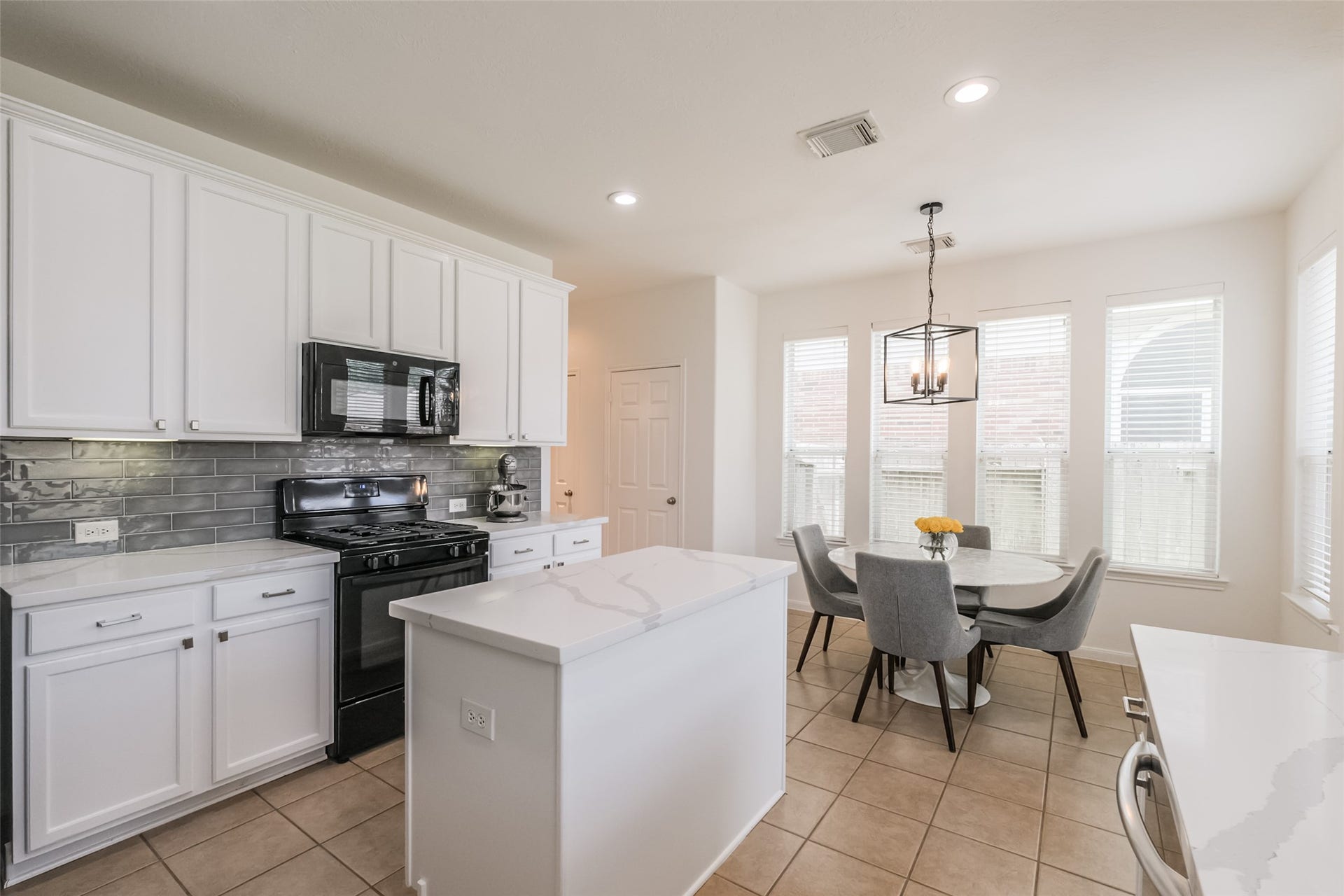 10314 Five Oaks Crossing, Missouri City, TX 77459 This modern kitchen features sleek white cabinetry, a stylish gray backsplash, and a central island with ample countertop space. It includes a black stove and microwave, with a cozy dining area bathed in natural light from large windows. A contemporary light fixture adds a touch of elegance.