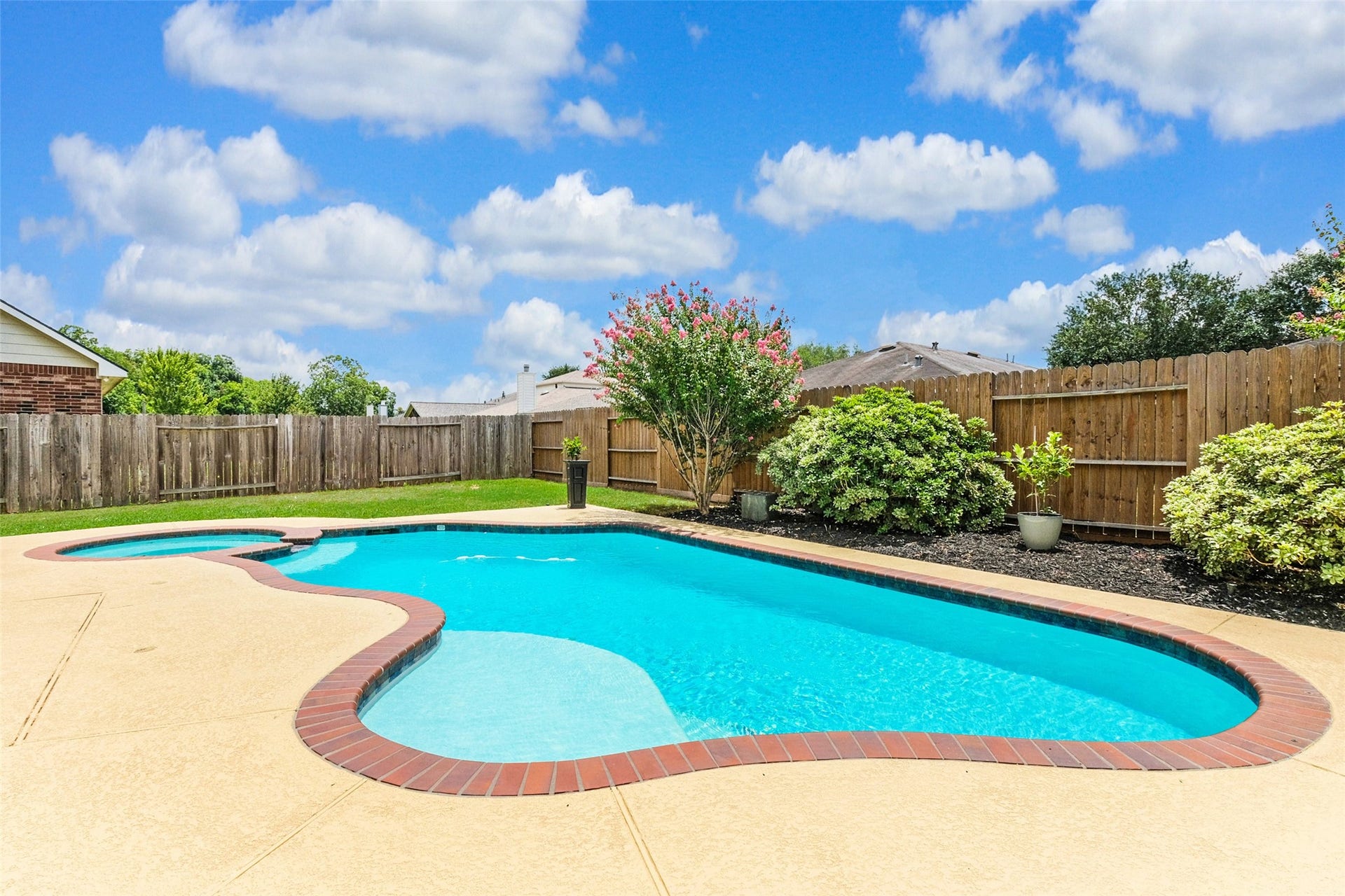 10314 Five Oaks Crossing, Missouri City, TX 77459 This photo shows a backyard with a well-maintained, curvy swimming pool surrounded by a concrete deck. The yard is enclosed by a wooden fence and features neatly landscaped greenery, creating a private and inviting outdoor space.