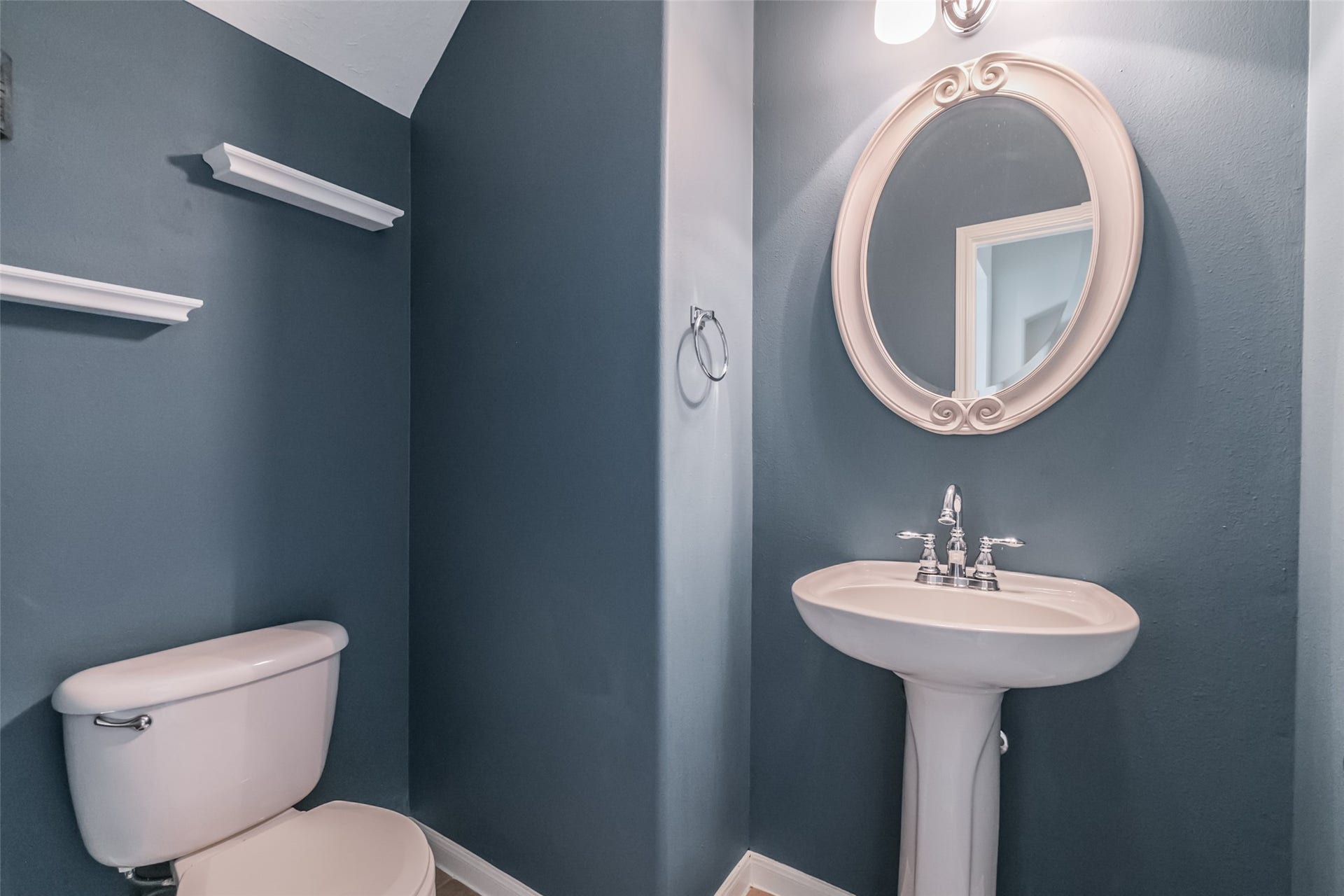 10314 Five Oaks Crossing, Missouri City, TX 77459 Compact half-bathroom with stylish gray-blue walls, featuring a pedestal sink, oval mirror, and modern light fixture. Practical shelving and a sleek design complete the space.