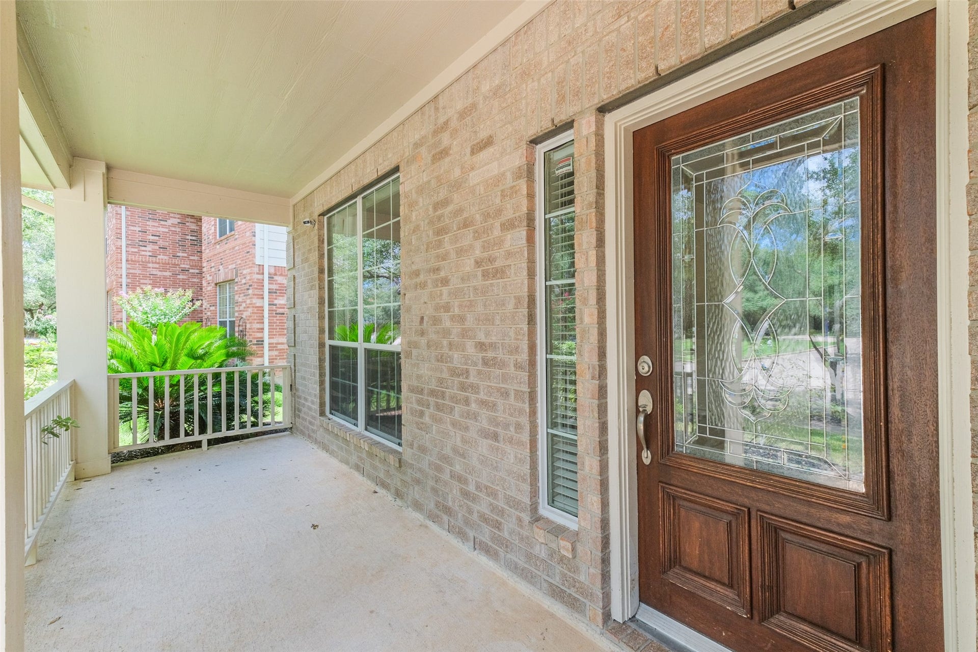 10314 Five Oaks Crossing, Missouri City, TX 77459 A welcoming front porch with a brick exterior features a decorative glass front door and large windows. The space is sheltered and overlooks a small garden with lush greenery, providing a charming entrance to the home.