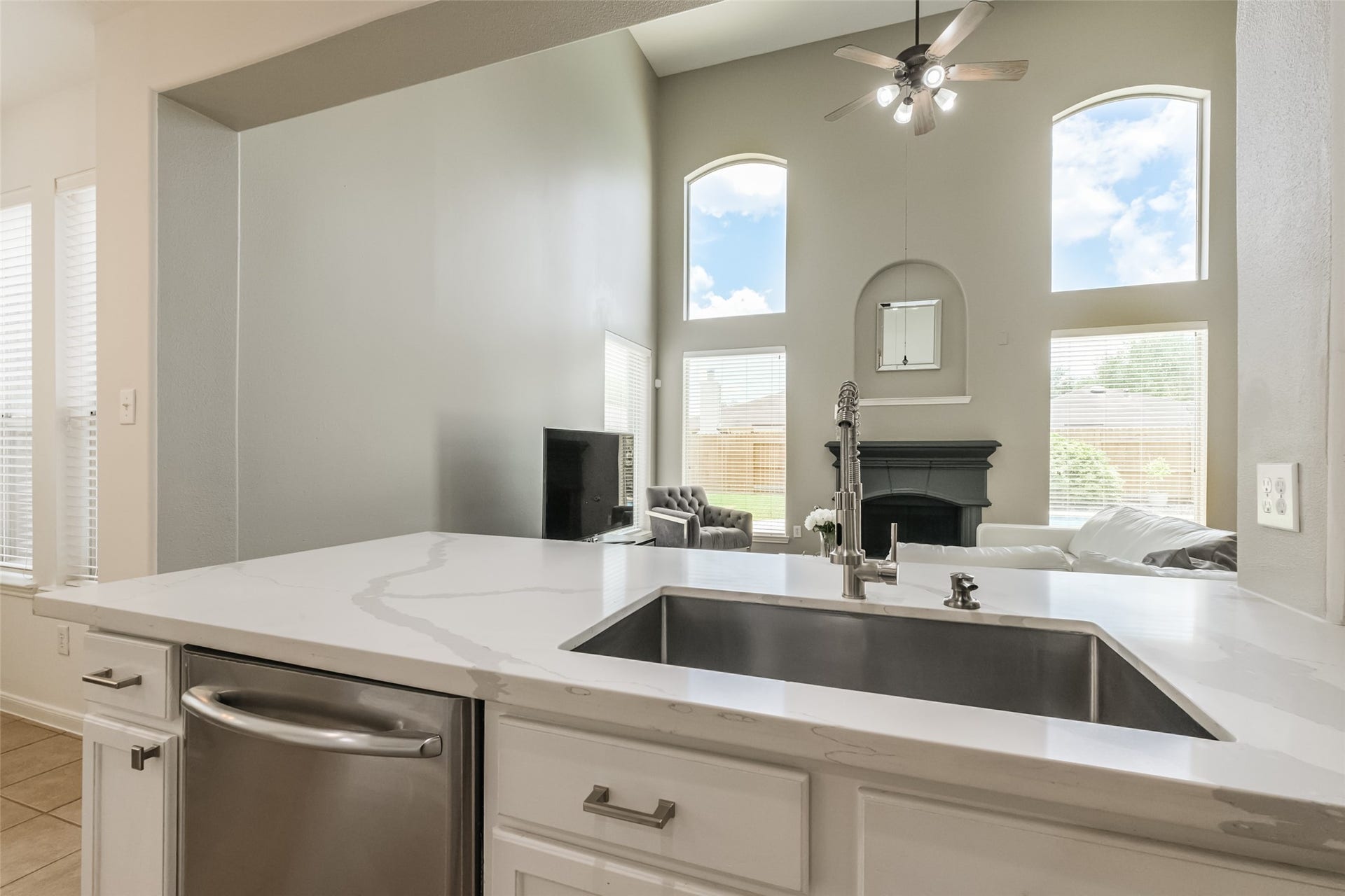 10314 Five Oaks Crossing, Missouri City, TX 77459 This photo showcases a modern open-concept living area with high ceilings and large windows, providing plenty of natural light. The view from the kitchen features a sleek countertop with a built-in sink, overlooking a cozy living room with a fireplace and ceiling fan, creating a bright and inviting space.
