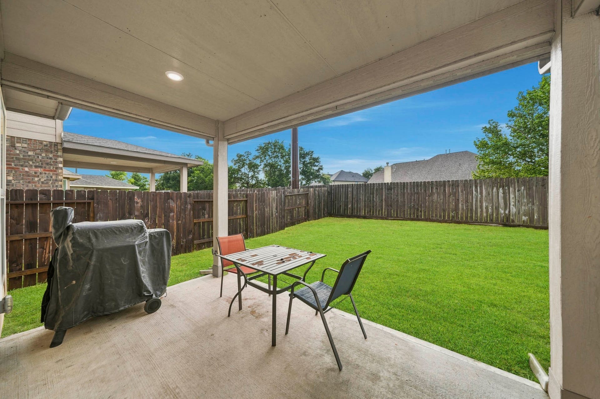 Covered patio with seating area and barbecue setup, overlooking a spacious, fenced backyard with lush green grassperfect for outdoor entertaining.