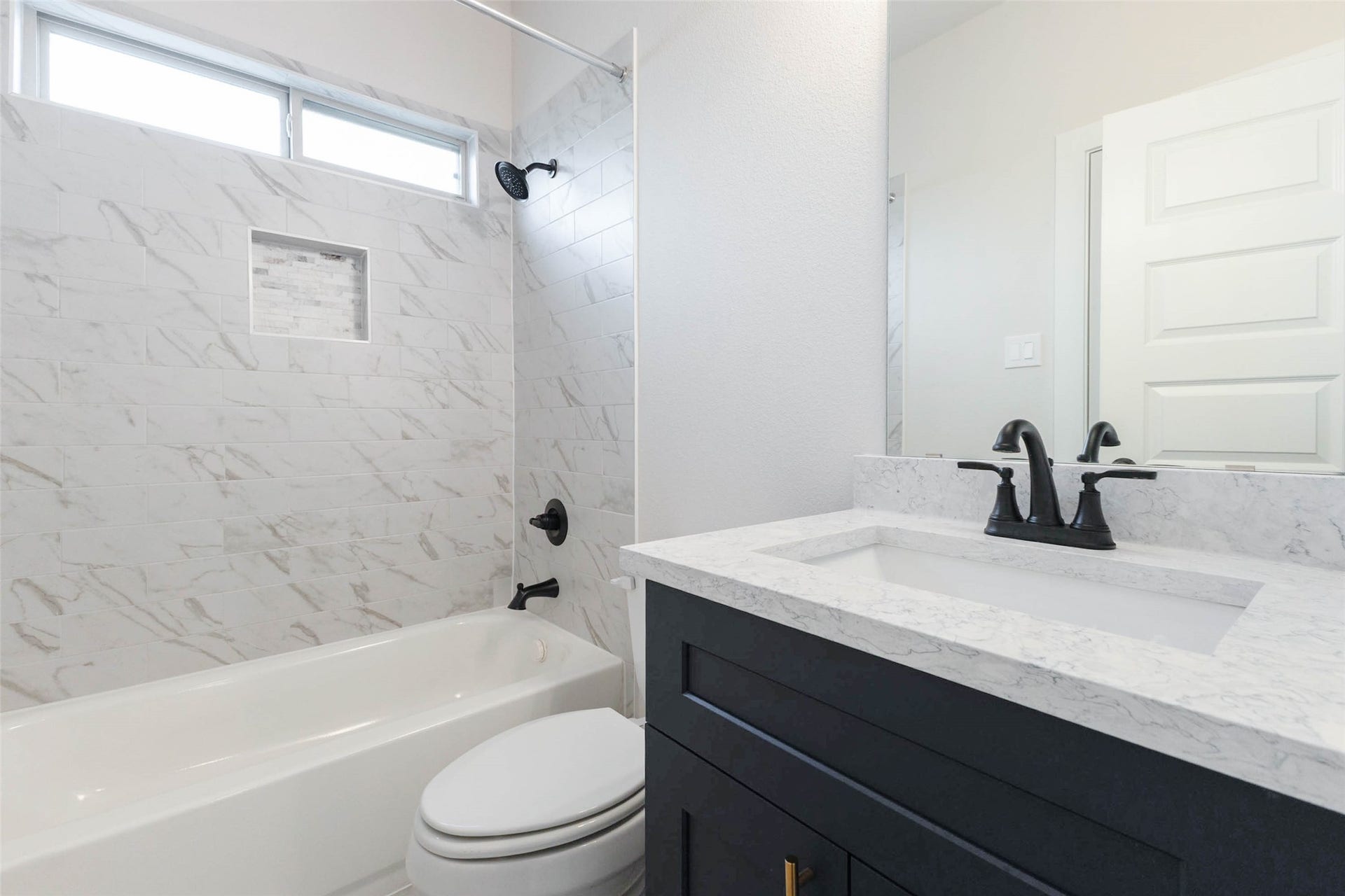 1007 Santee Ct Court, Rosharon, TX 77583 A full bath near the bedroom wing features a tub-shower wrapped in marble-look tile with a built-in niche for shampoo and soap. The navy vanity, quartz top and matte black fixtures create a modern, high-end look, and a transom window above the tub brings in natural light. It is a practical, stylish space for guests, kids or anyone needing a bath close to their bedroom.