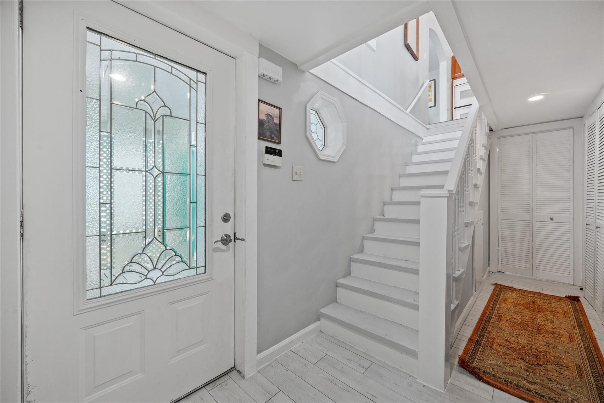 1805 19th Street, Galveston, TX 77550 Front Entrance & Staircase: Welcoming front entrance showcasing a beautiful decorative leaded-glass entry door and classic white staircase, highlighting the home's charming architectural character. Wood-look tile flooring and coastal-inspired finishes complement the inviting entryway, with stairs leading to the second-floor kitchen, living and dining areas, and outdoor upper deck.