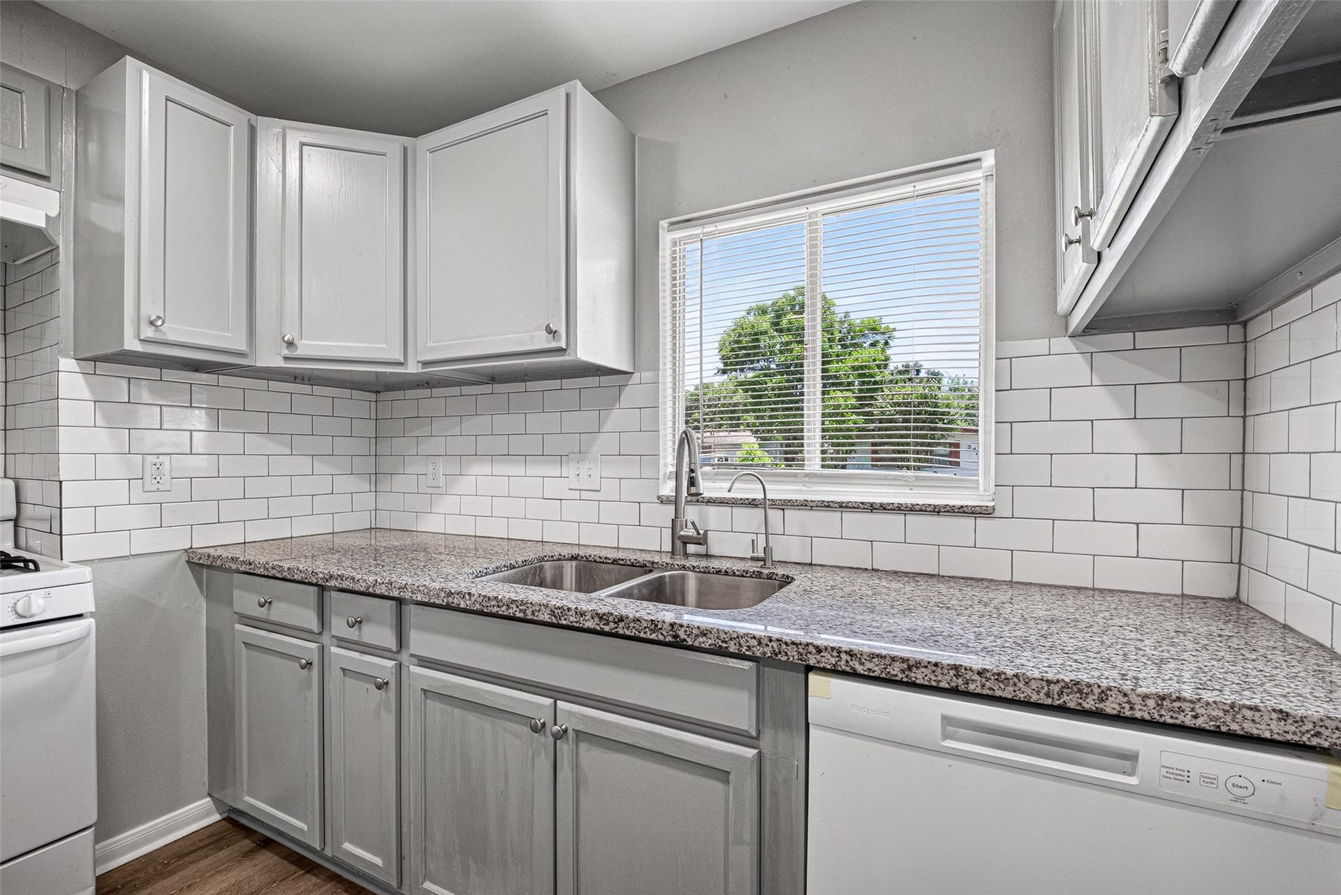 3408 Palm Avenue, Texas City, TX 77590 This kitchen features sleek granite countertops, white cabinetry, and a modern subway tile backsplash. It includes a double sink, dishwasher, and a window with a view, offering a bright and functional space.