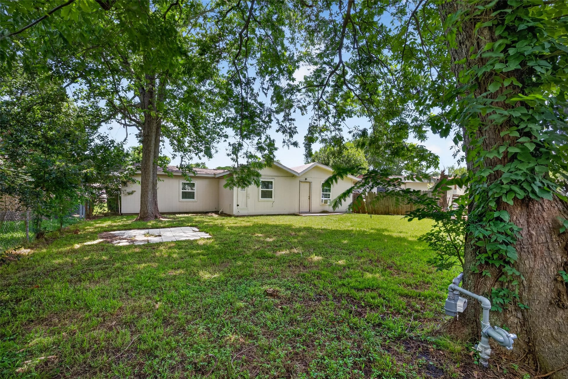 3408 Palm Avenue, Texas City, TX 77590 This photo shows a cozy backyard with mature trees providing shade, a grassy area, and a charming single-story home with light-colored siding. It's a peaceful, private space perfect for relaxing or entertaining.