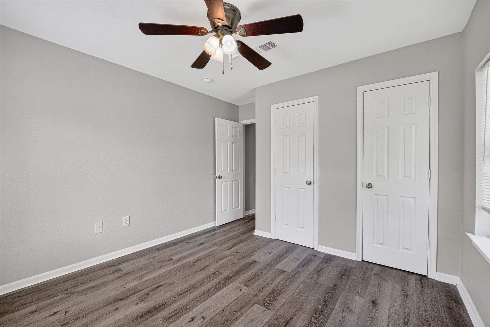 3408 Palm Avenue, Texas City, TX 77590 This room features modern gray walls, wood-look flooring, a ceiling fan with lights, and ample natural light from a window. It includes two white doors, likely for closet and entry access, creating a clean and functional space.