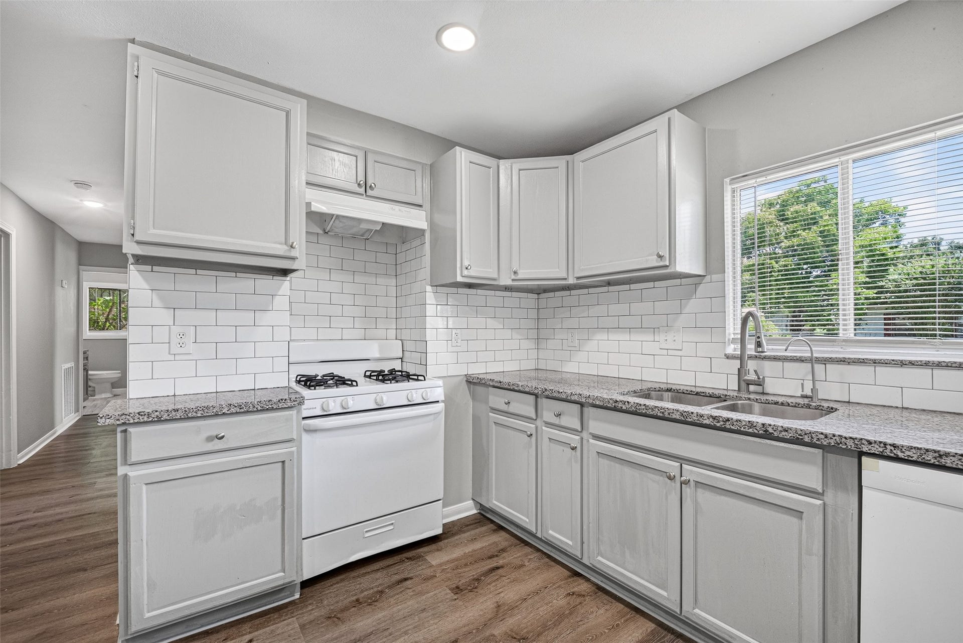 3408 Palm Avenue, Texas City, TX 77590 This kitchen features modern gray cabinetry, granite countertops, and a classic subway tile backsplash. It includes a gas stove and a double sink under a bright window, complemented by wood-style flooring.