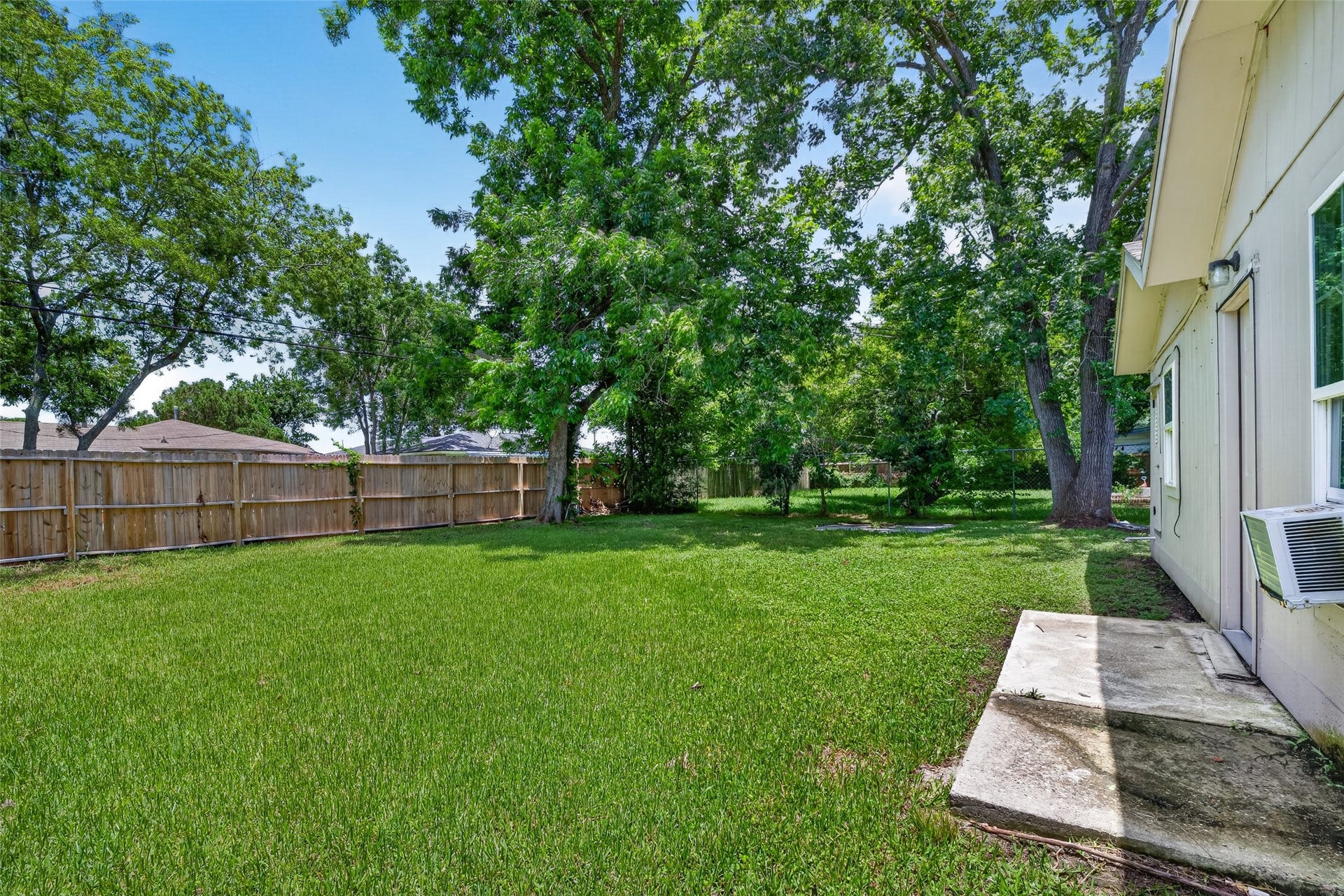 3408 Palm Avenue, Texas City, TX 77590 This photo shows a spacious backyard with lush green grass and mature trees, enclosed by a wooden privacy fence. The side of the house, with a window and exterior door, is partially visible. Ideal for outdoor activities and relaxation.