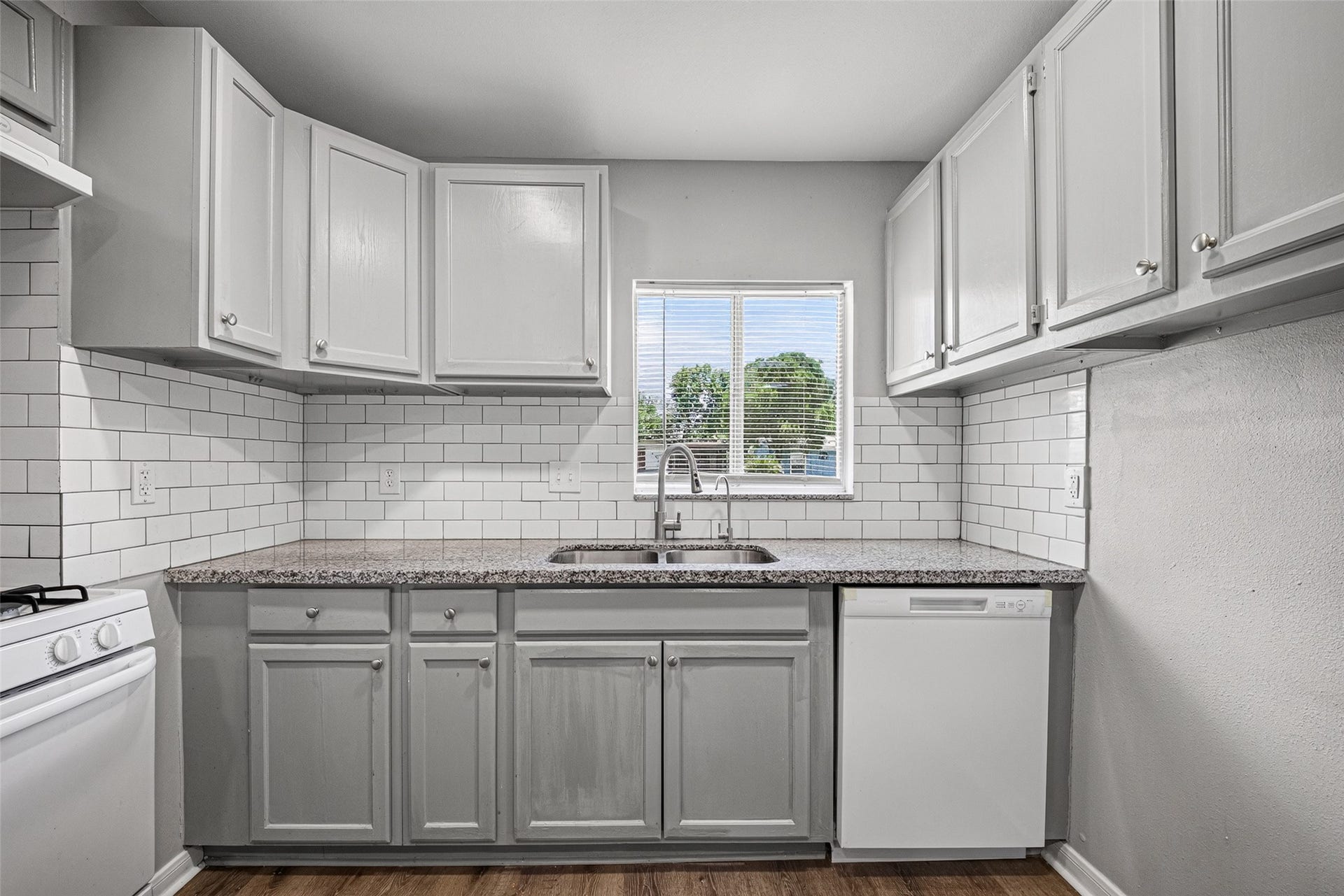 3408 Palm Avenue, Texas City, TX 77590 This kitchen features modern, light gray cabinets with sleek hardware, a granite countertop, and a subway tile backsplash. It includes a window above the sink for natural light, a dishwasher, and a gas stove, all complemented by wood-style flooring.