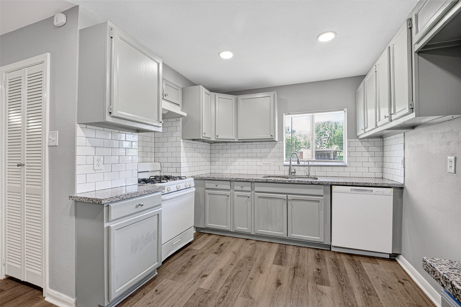 3408 Palm Avenue, Texas City, TX 77590 This kitchen features a modern, clean design with light gray cabinets, granite countertops, and white subway tile backsplash. It includes a gas stove, dishwasher, and a window above the sink for natural light. The space is complemented by wood-style flooring and recessed lighting.