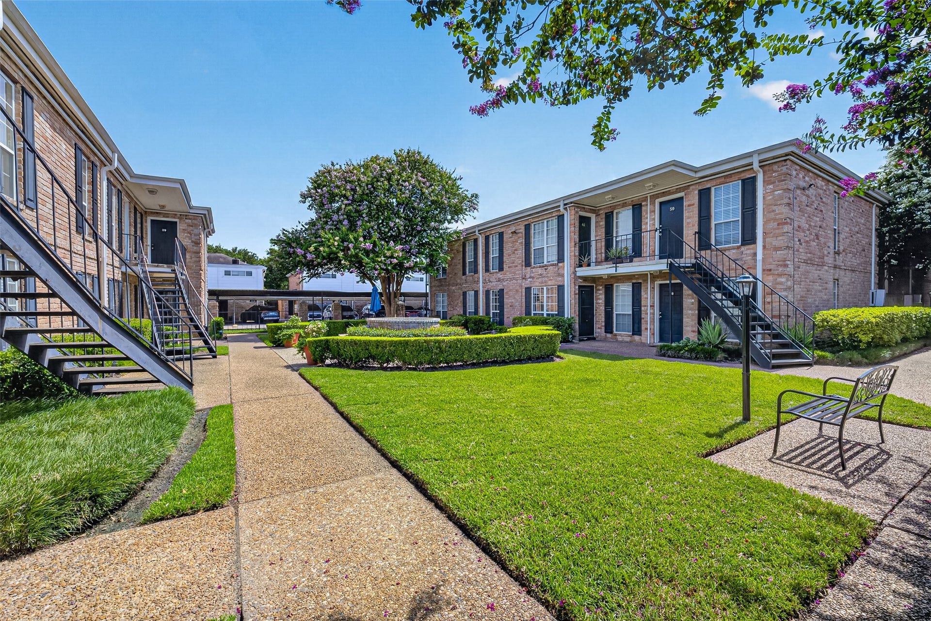 6402 Del Monte Drive, #65, Houston, TX 77057 This is a photo of a charming, well-maintained apartment complex featuring two-story brick buildings with external staircases. The landscaped courtyard includes lush green lawns, manicured hedges, and a mature tree. A pathway leads through the area, offering a pleasant, inviting atmosphere.