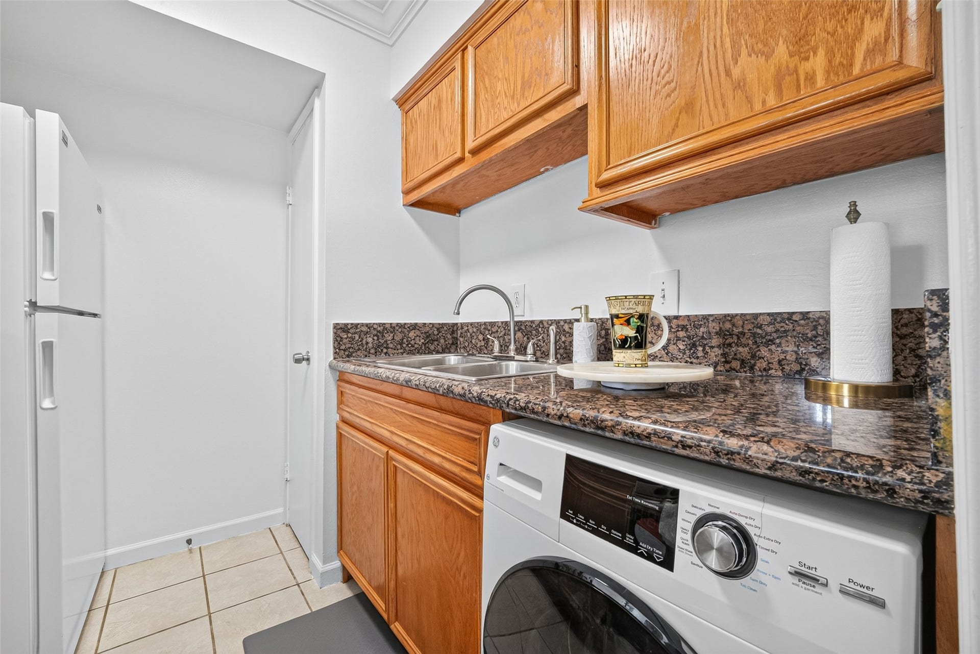 6402 Del Monte Drive, #65, Houston, TX 77057 This photo shows a compact laundry area with a washing machine, granite countertops, and wooden cabinets. There's a stainless steel sink and a small workspace, creating a functional and efficient space.