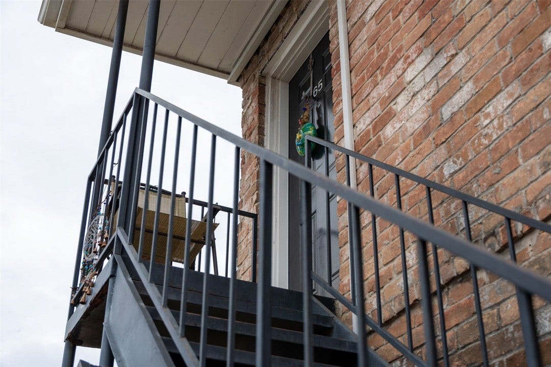 6402 Del Monte Drive, #65, Houston, TX 77057 This photo shows a brick exterior with a metal staircase leading to a second-floor entrance. The area appears to have a small balcony space with a chair.