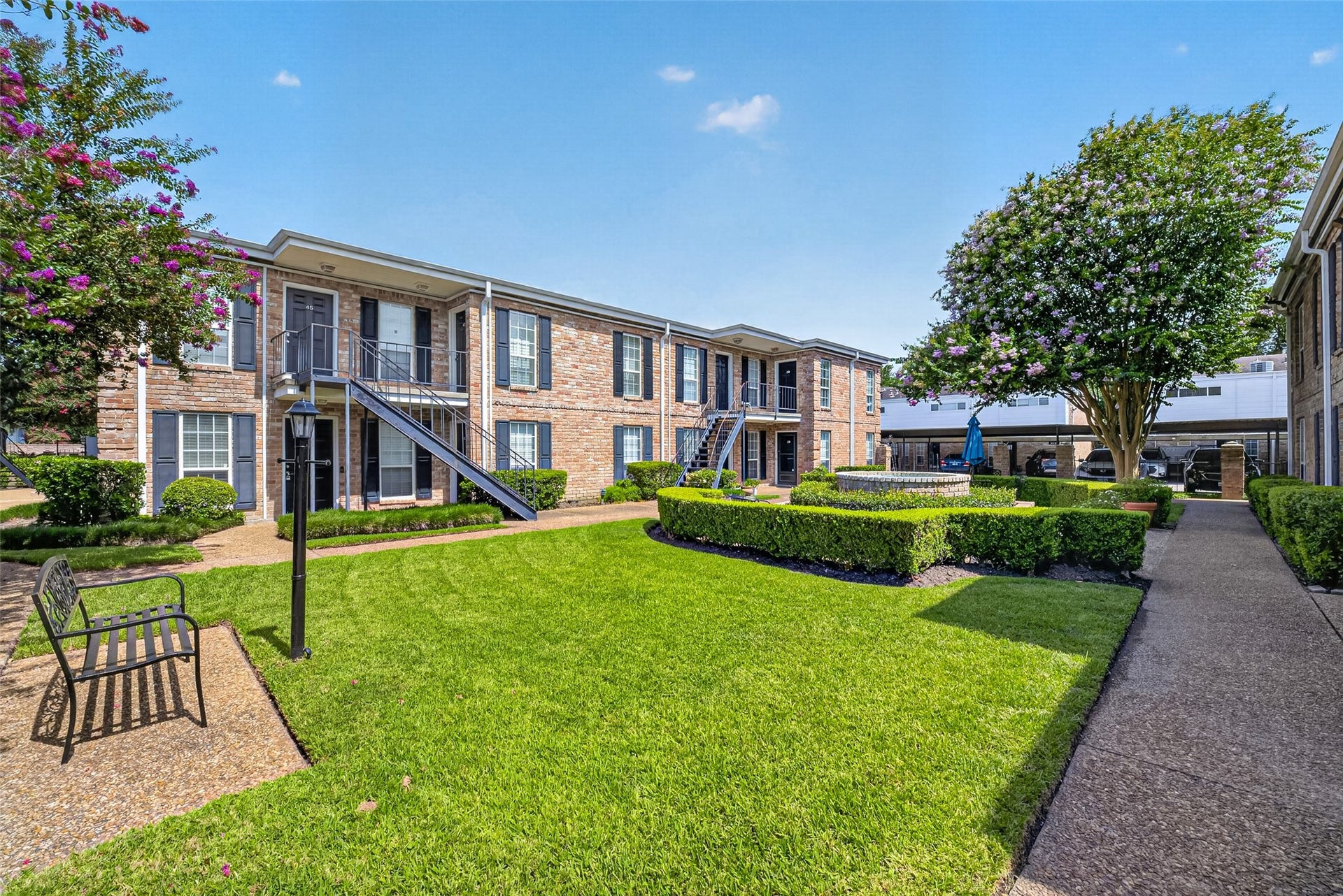 6402 Del Monte Drive, #65, Houston, TX 77057 This photo showcases a charming apartment complex with well-maintained landscaping, featuring lush green lawns, neatly trimmed hedges, and mature flowering trees. The two-story brick buildings have exterior staircases, and a central courtyard offers a peaceful seating area.