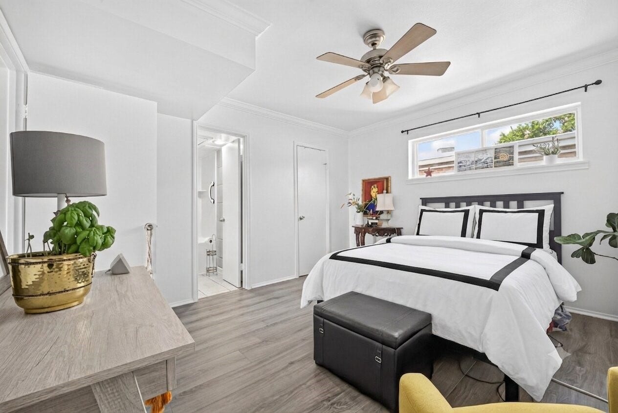 6402 Del Monte Drive, #65, Houston, TX 77057 This bedroom features a modern, bright design with white walls and wood flooring. It includes a cozy bed with black and white linens, a ceiling fan, and a small window for natural light. The room is complemented by a wooden desk, decorative plants, and an en-suite bathroom.