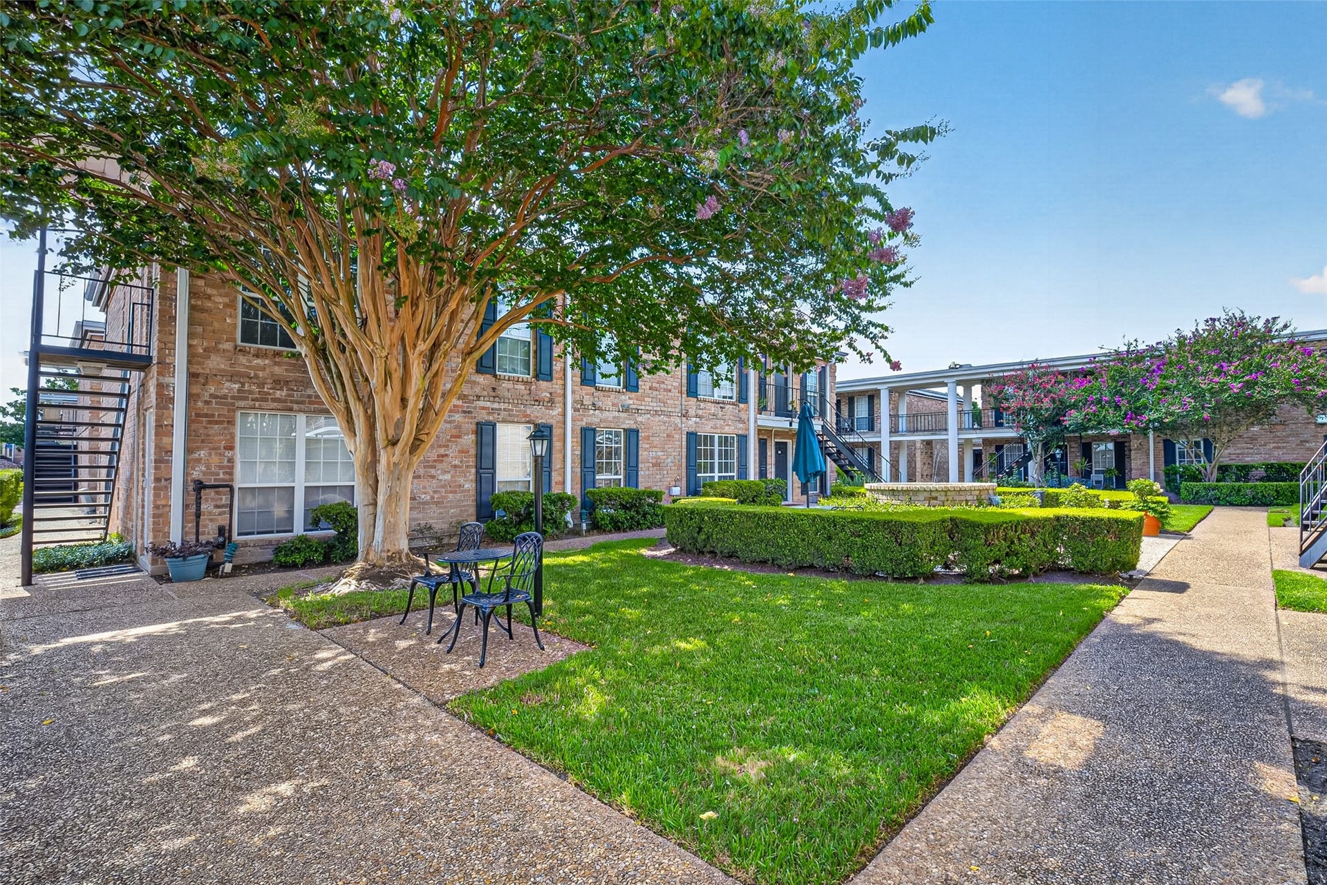 6402 Del Monte Drive, #65, Houston, TX 77057 This photo showcases a charming courtyard in an apartment complex, featuring a well-maintained lawn, mature trees, and a seating area. The brick building has two stories with external staircases, creating a welcoming and community-oriented atmosphere.